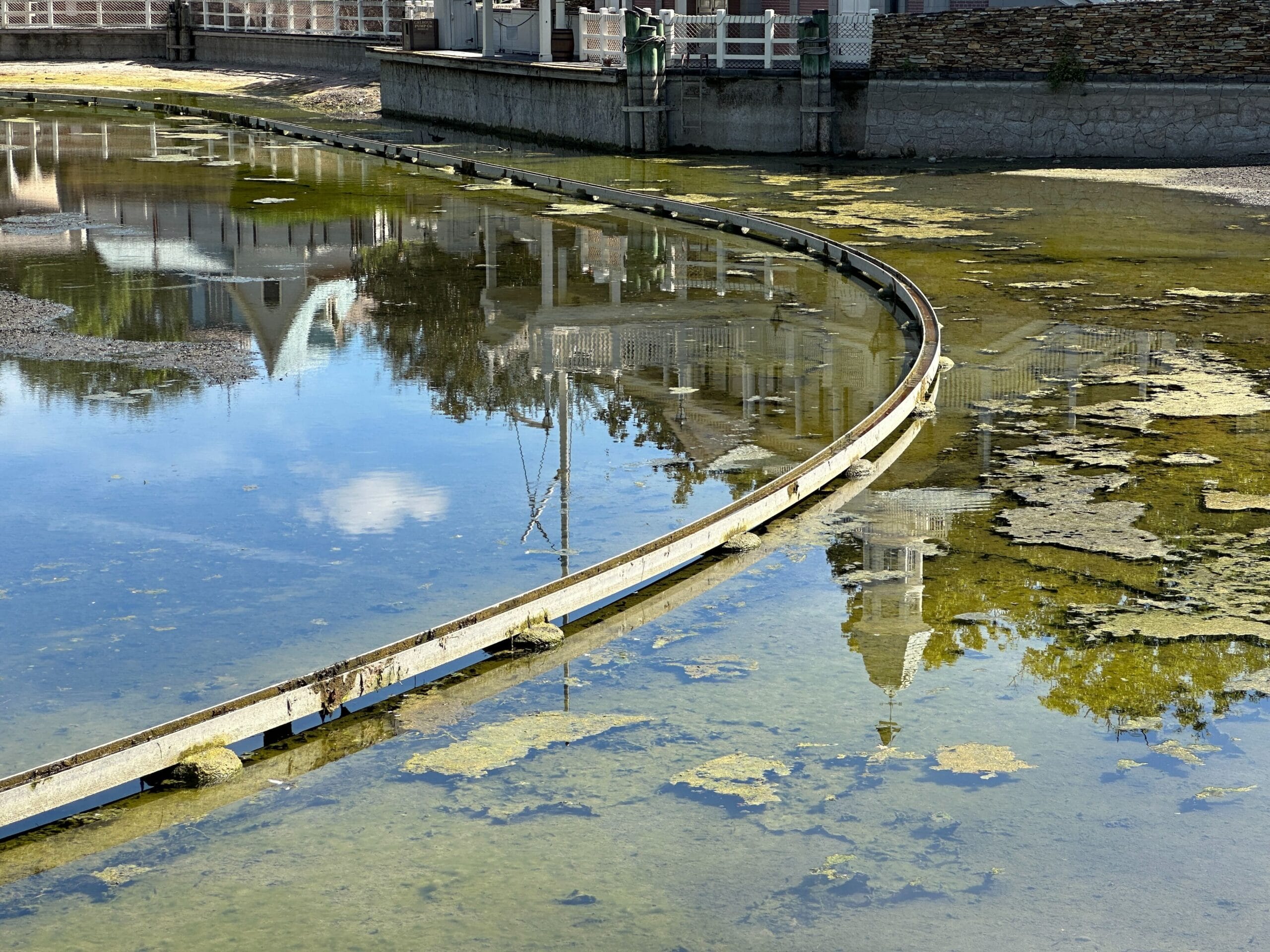 A curved metal barrier divides shallow, algae-filled water with building reflections under a sunny sky.