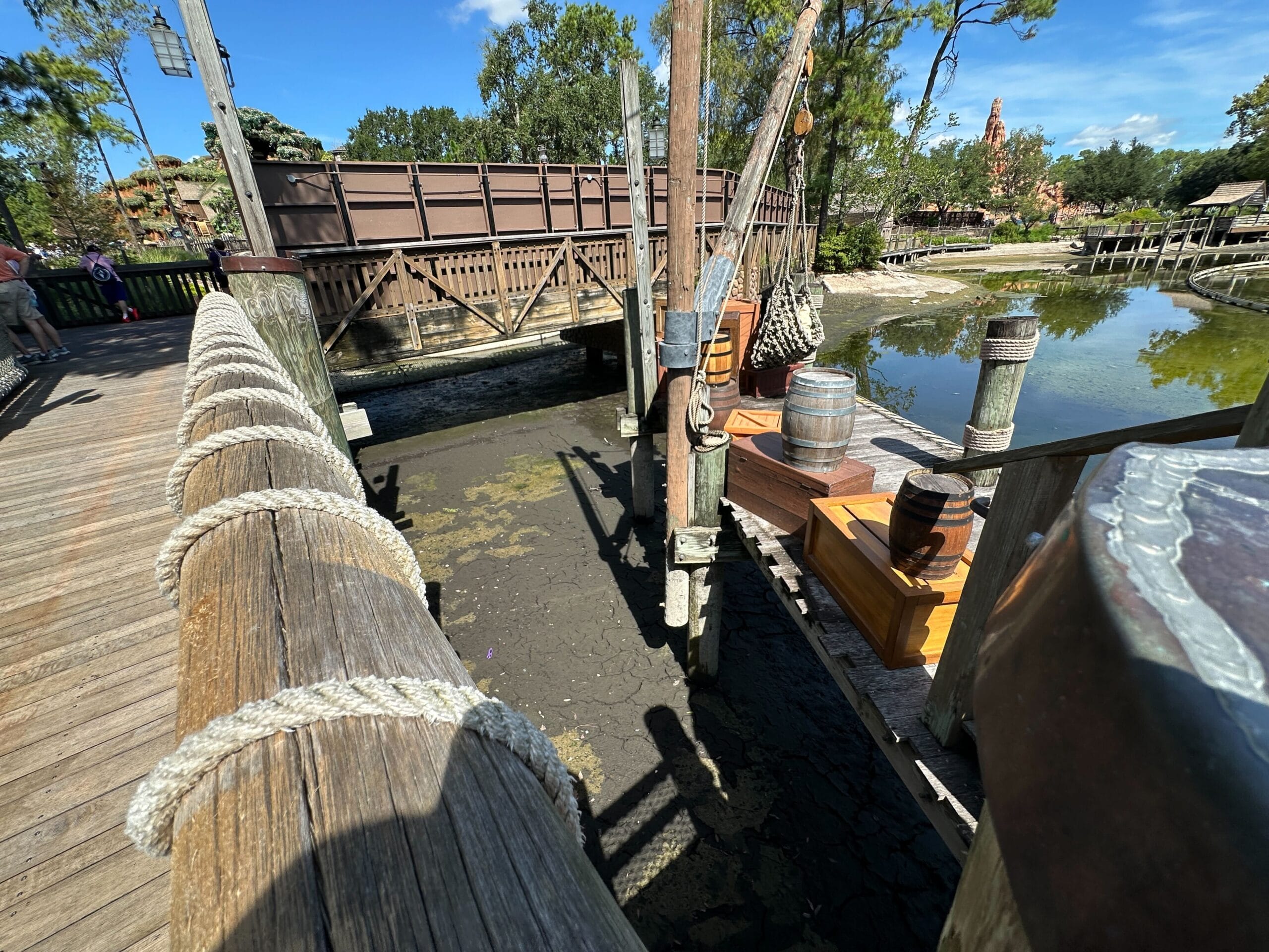 Wooden bridge over a muddy, low-water area with barrels and crates, trees and blue sky in the background.