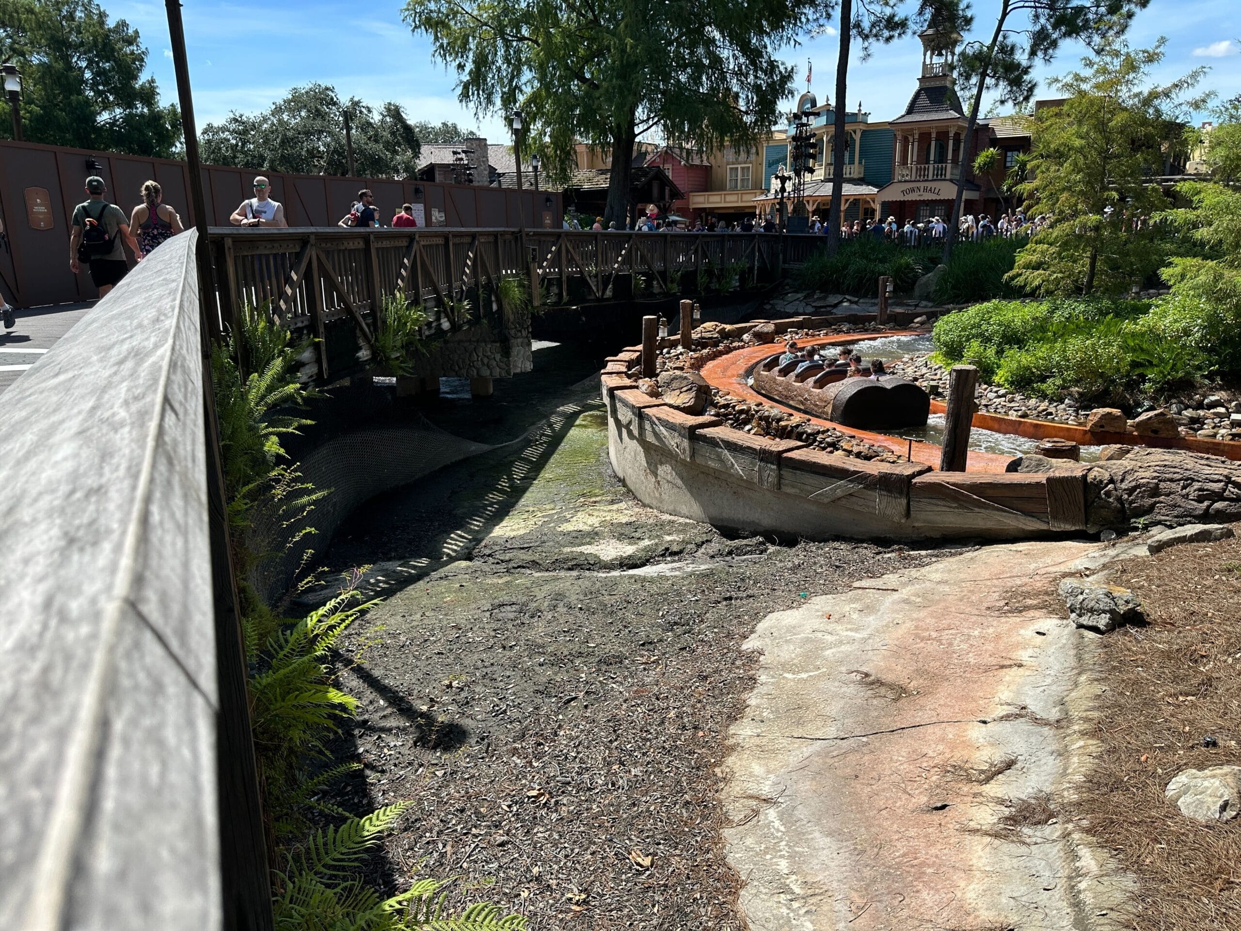 A dry water ride track with log boats, surrounded by plants, near a wooden bridge and people walking above.