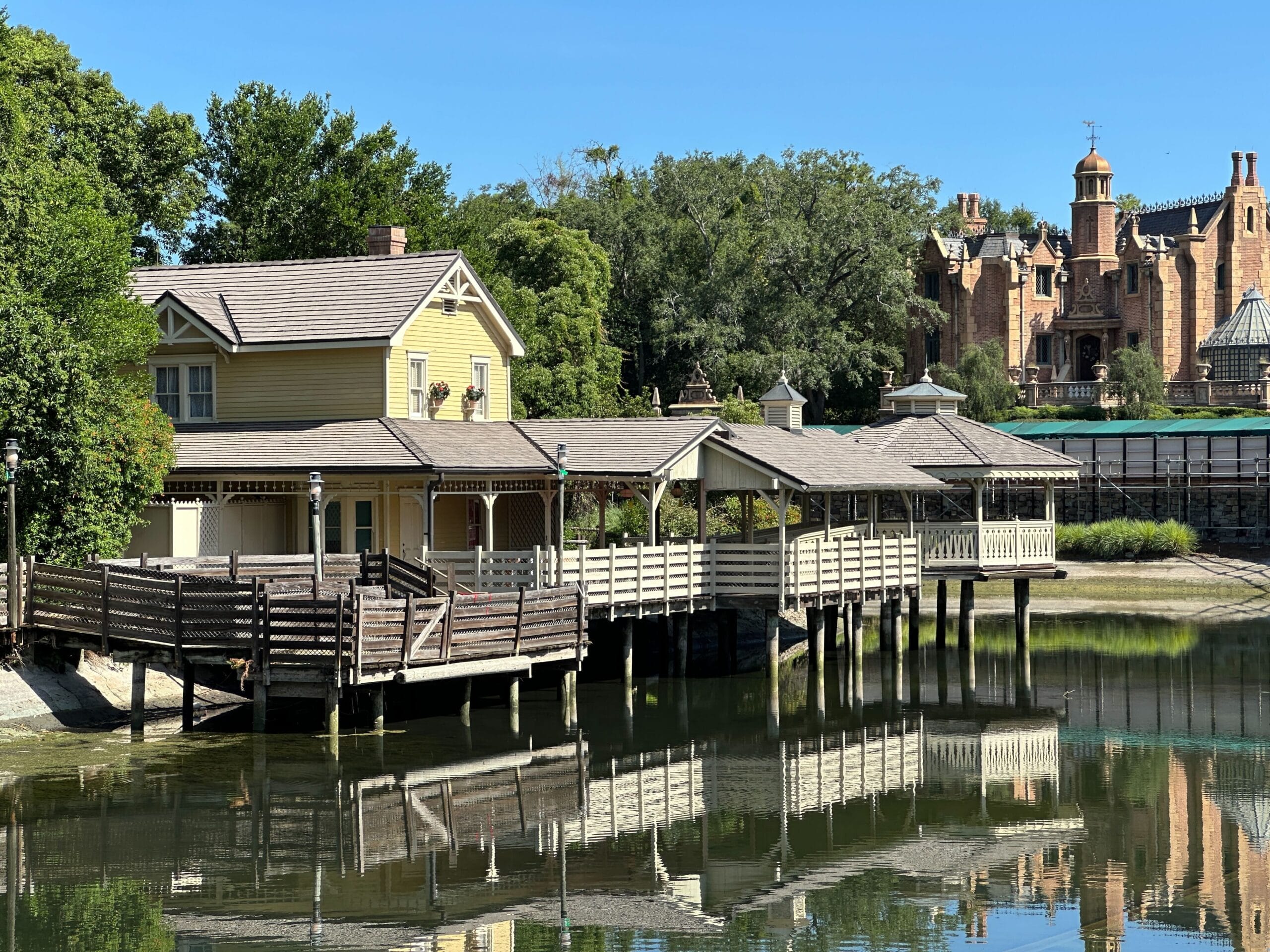 A wooden house and covered dock sit on a calm lake, with a large brick mansion in the background.