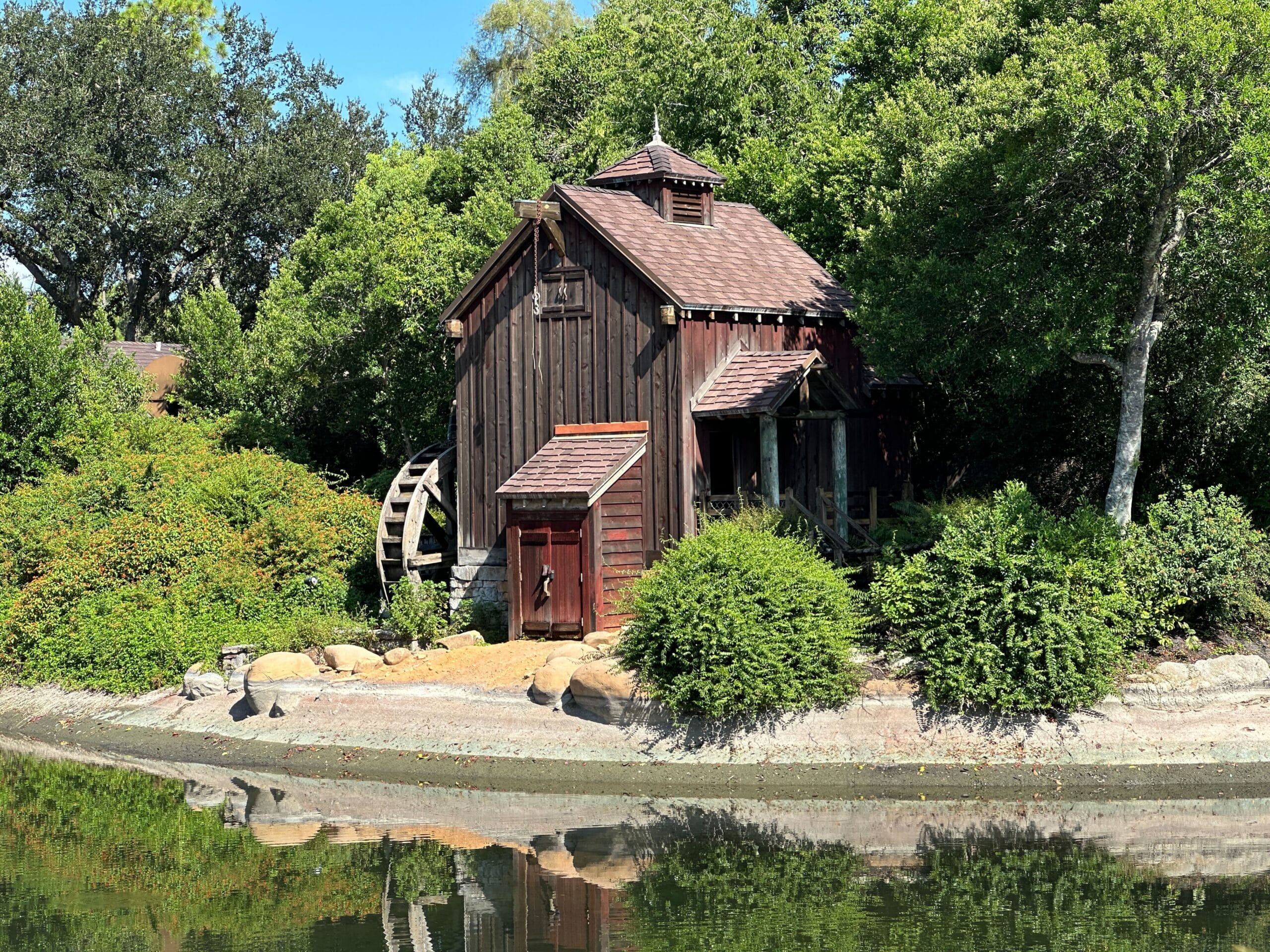 A rustic wooden watermill sits by a pond, surrounded by green trees and bushes under a blue sky.
