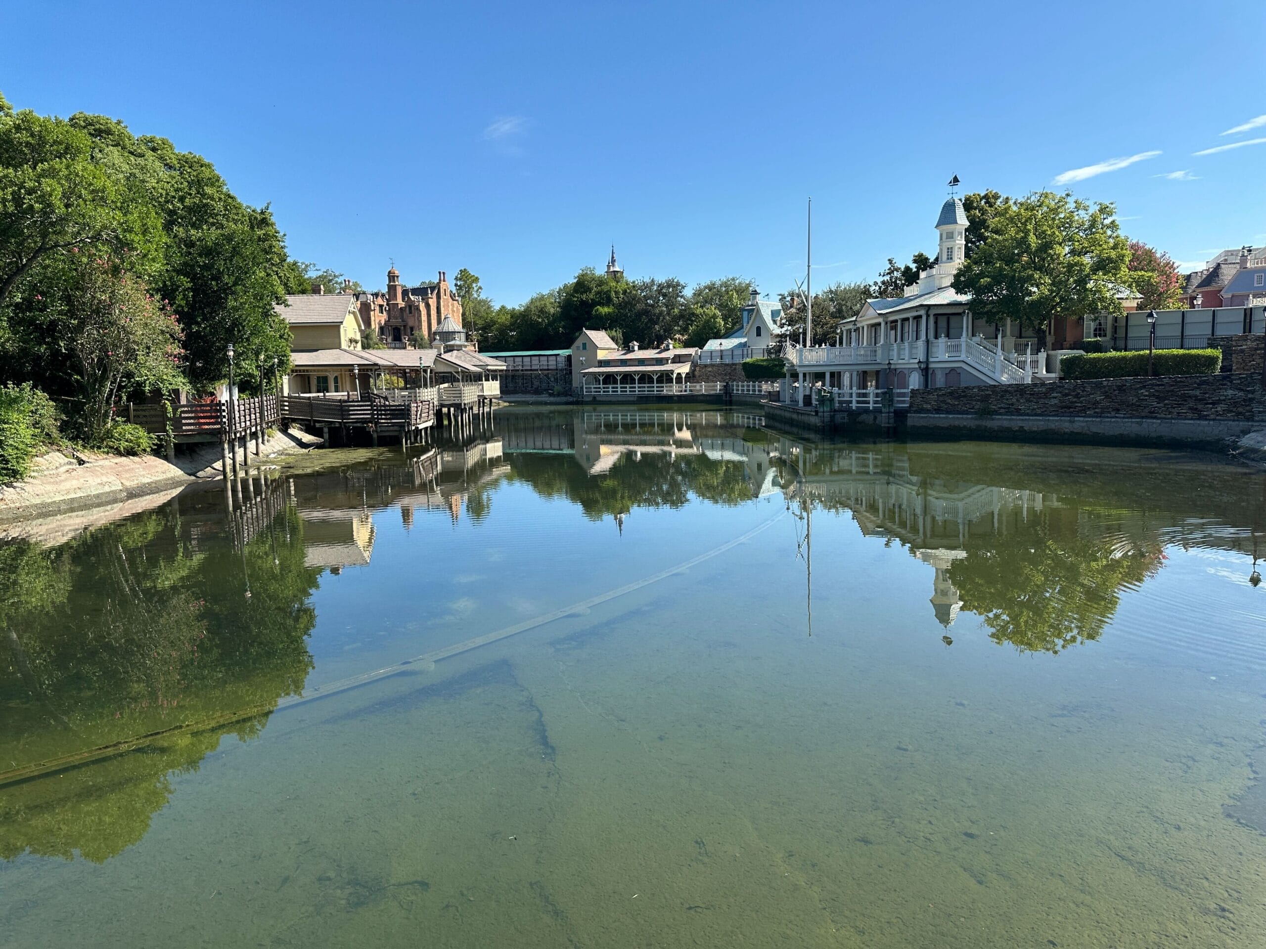 A clear, shallow river reflecting buildings and trees under a bright blue sky in a peaceful village setting.