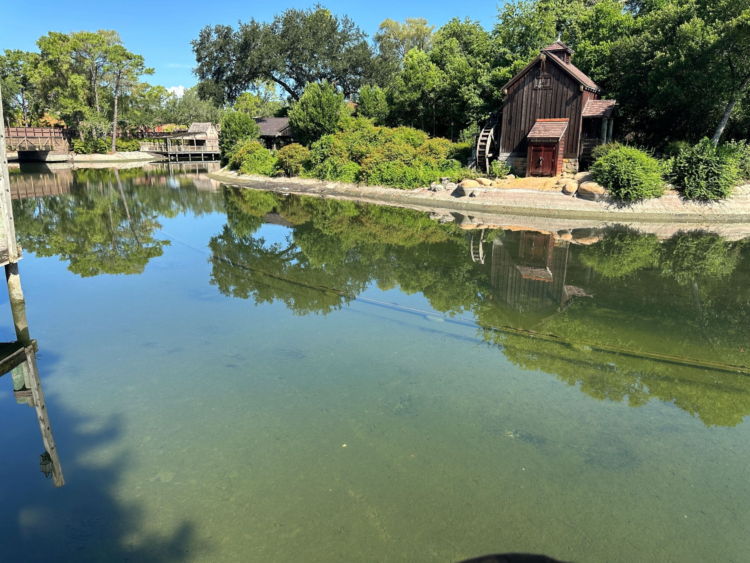 A rustic wooden cabin sits by a calm pond surrounded by trees and greenery under a clear blue sky.
