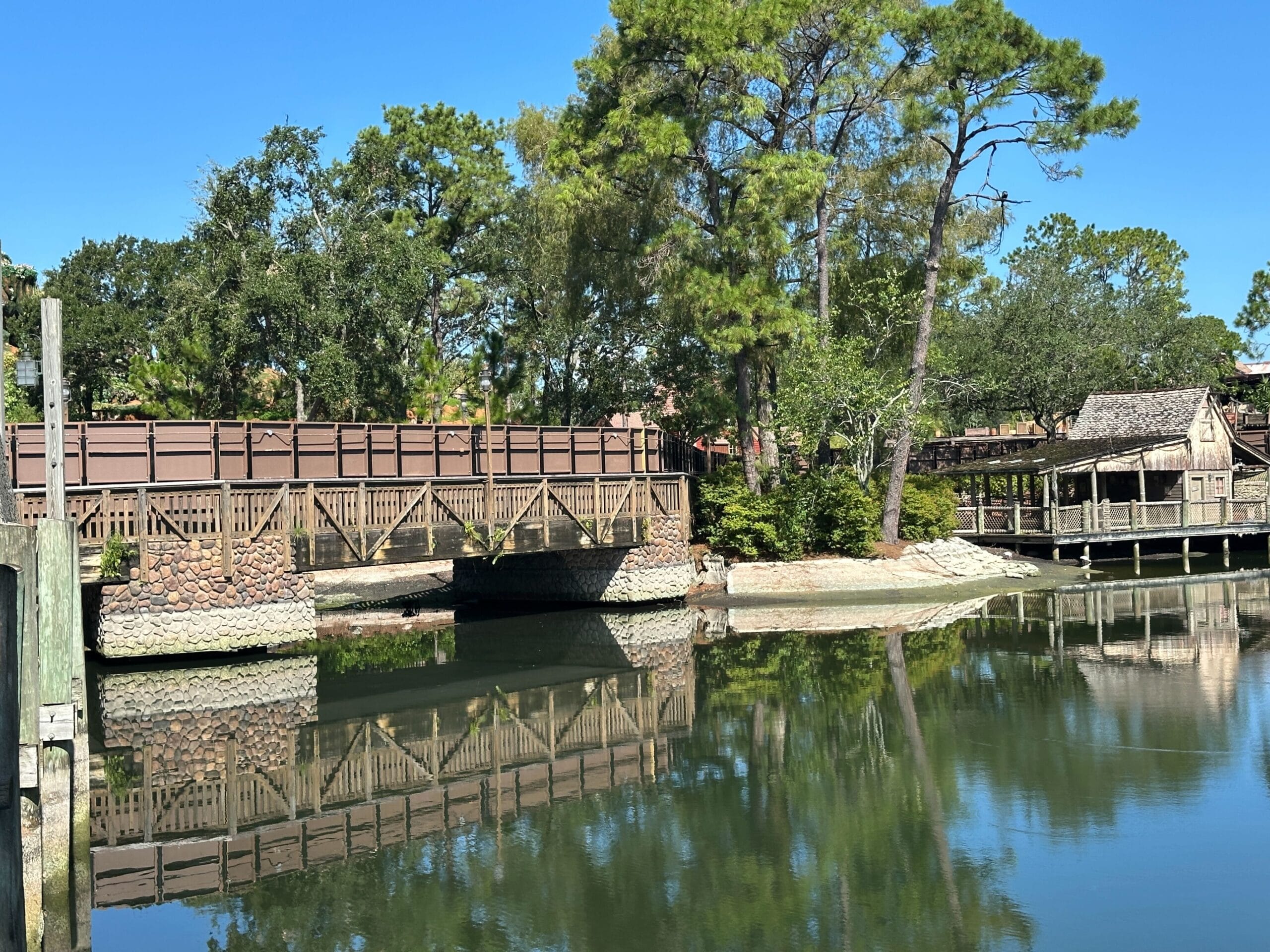 Wooden pedestrian bridge and rustic building reflected in calm water, surrounded by green trees under a clear blue sky.