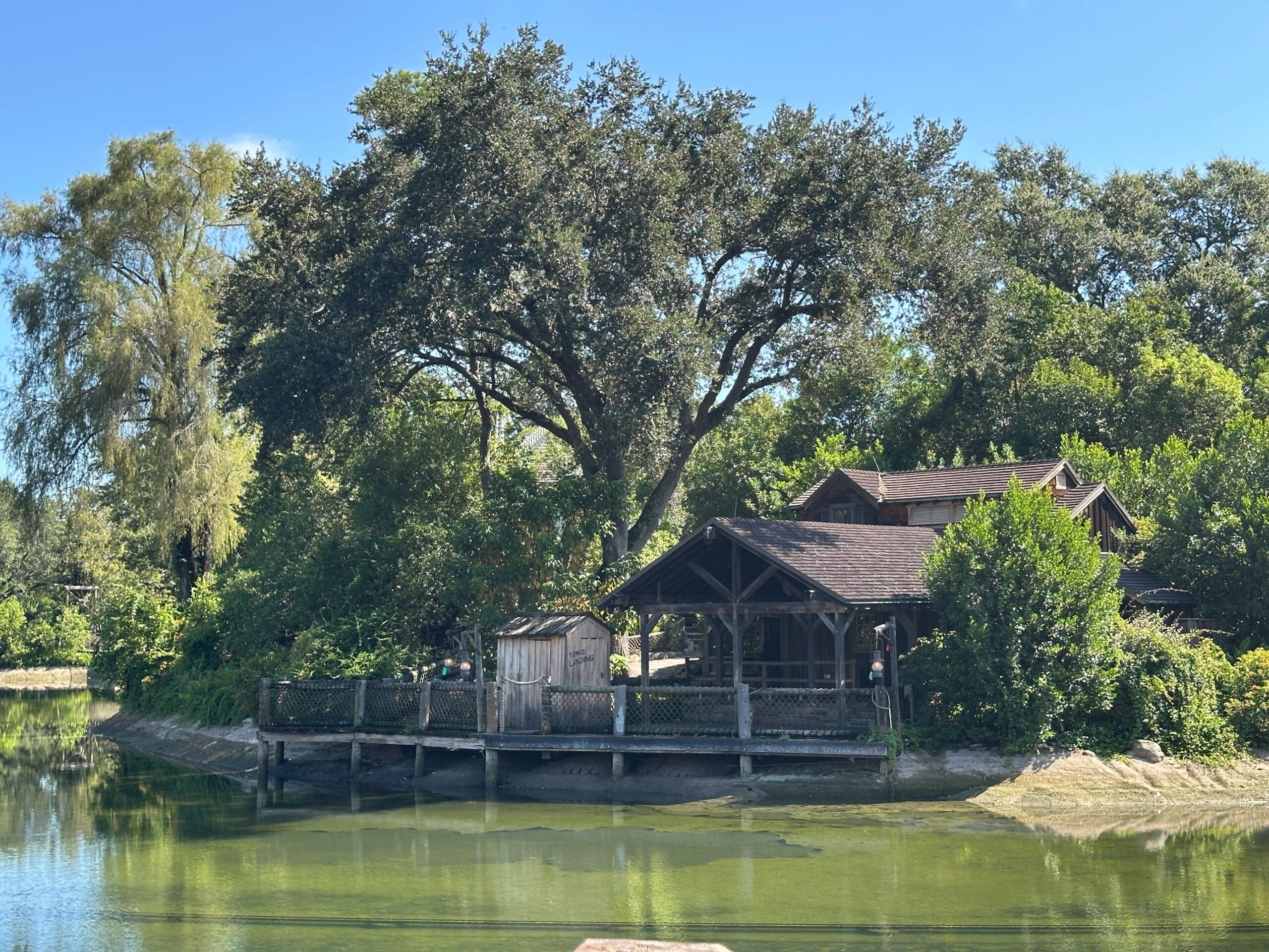 A rustic wooden cabin sits by a calm river, surrounded by trees and lush greenery under a clear blue sky.