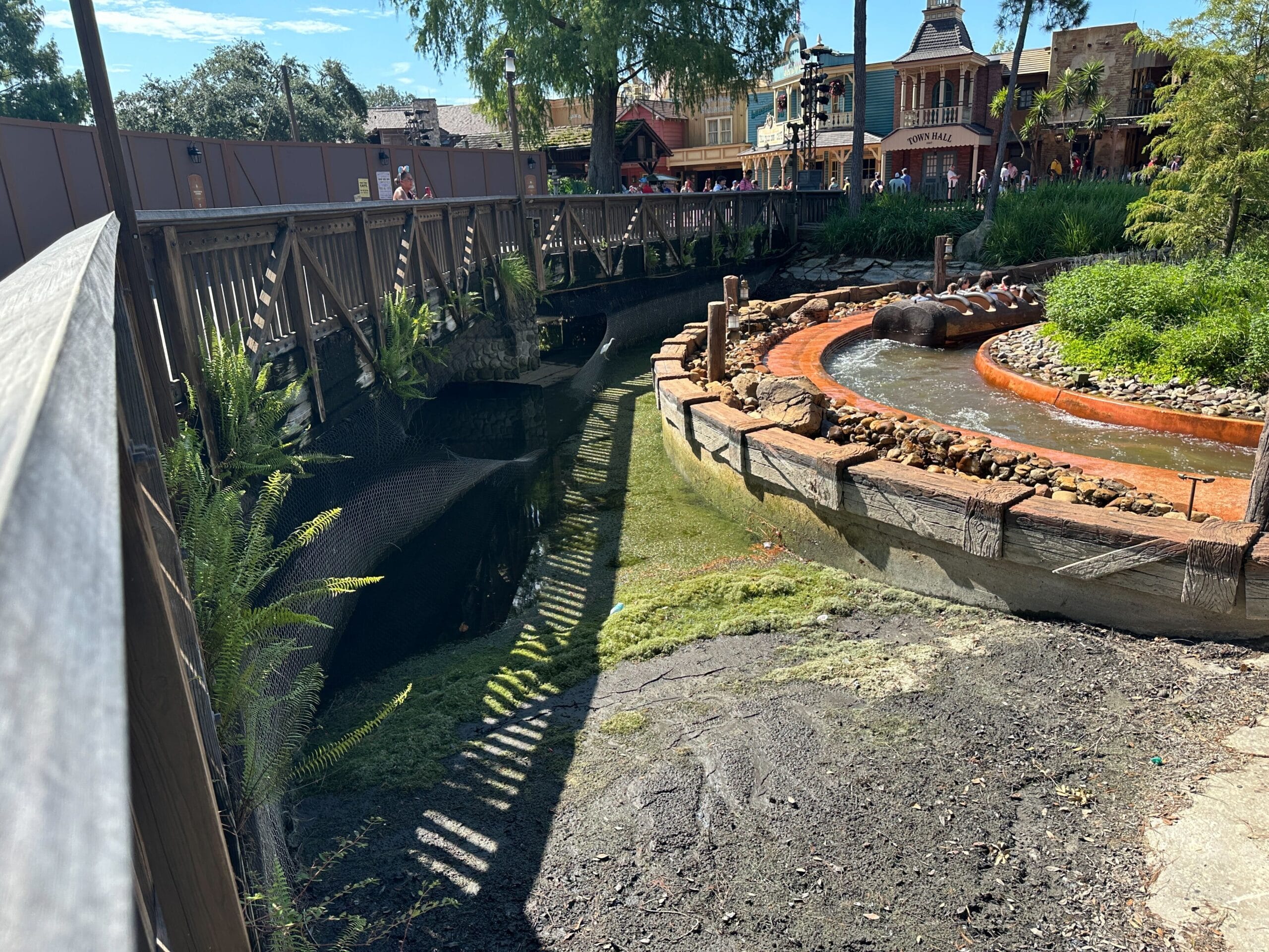 A dry, partially drained theme park waterway with a wooden bridge and construction barriers nearby under a sunny sky.