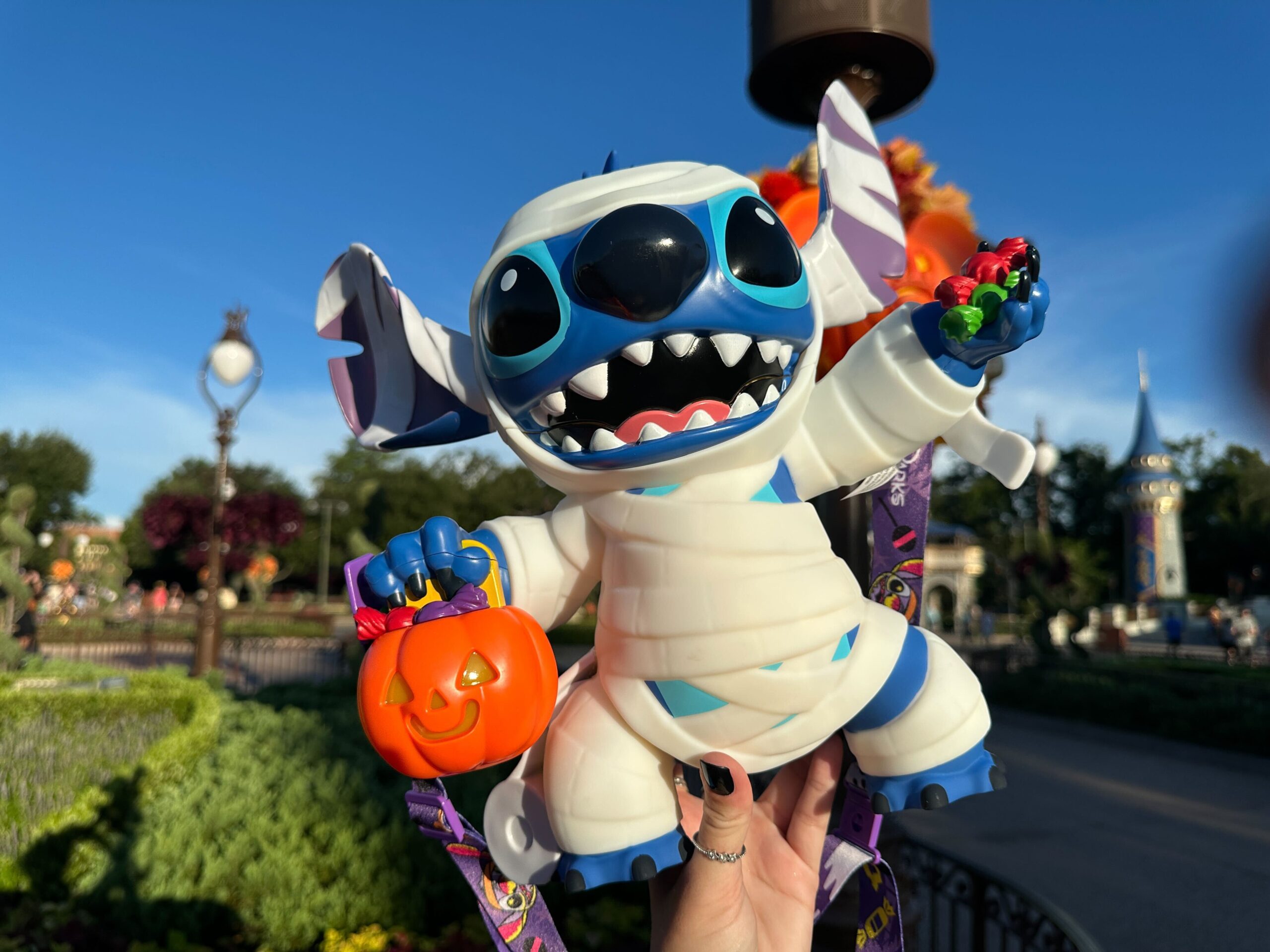A person holds a Stitch figure dressed as a mummy with a pumpkin bucket at a theme park on a sunny day.