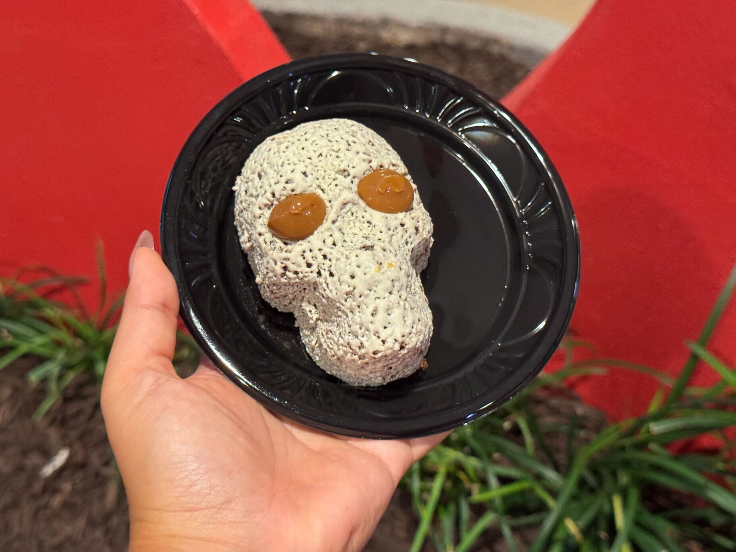 A hand holds a black plate with a skull-shaped dessert—perfect for food reviews from Mickey's Not So Scary Halloween Party.