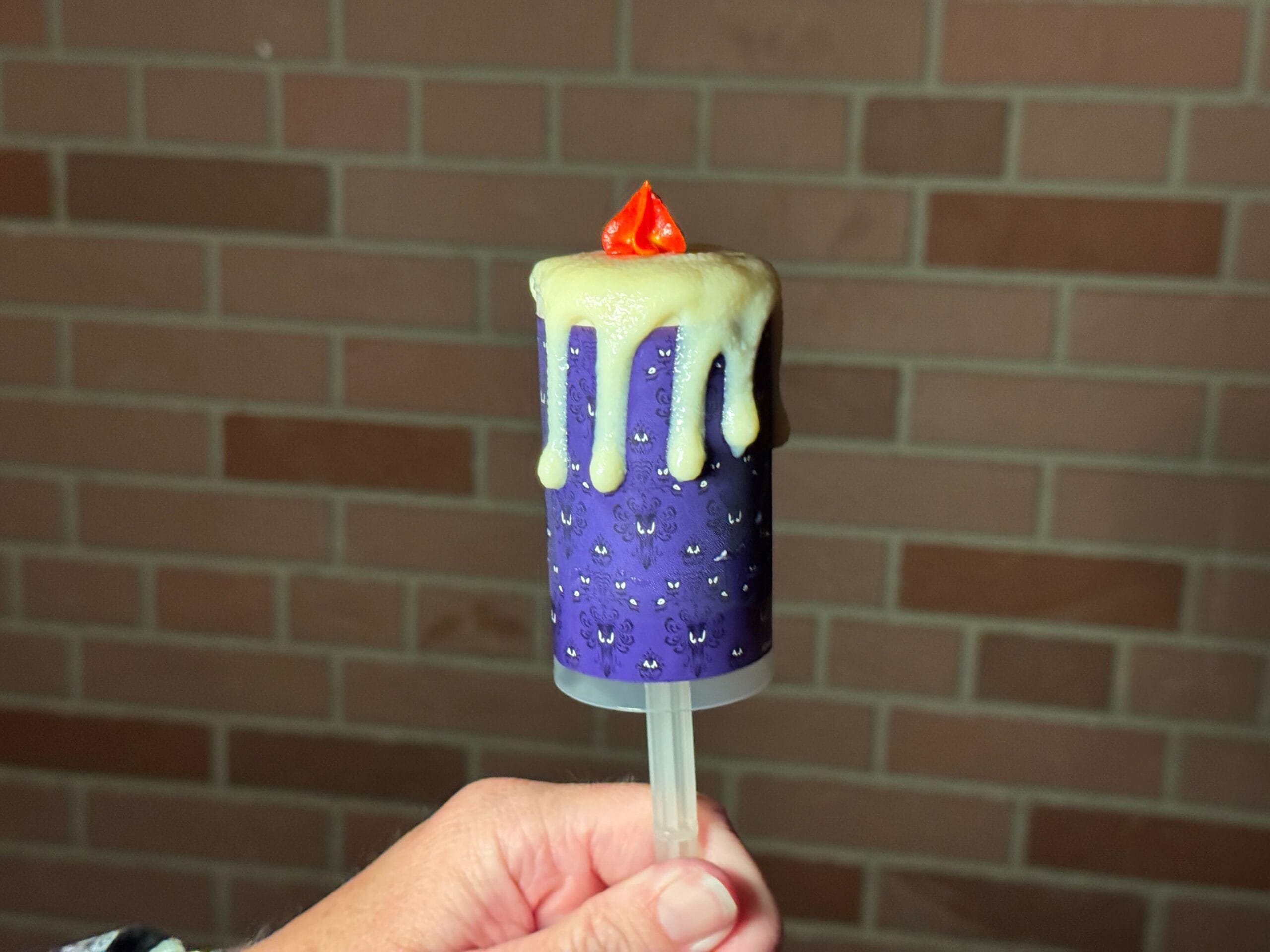 A hand holds a purple candle-shaped push pop at Mickey's Not So Scary Halloween Party, brick wall in the background.