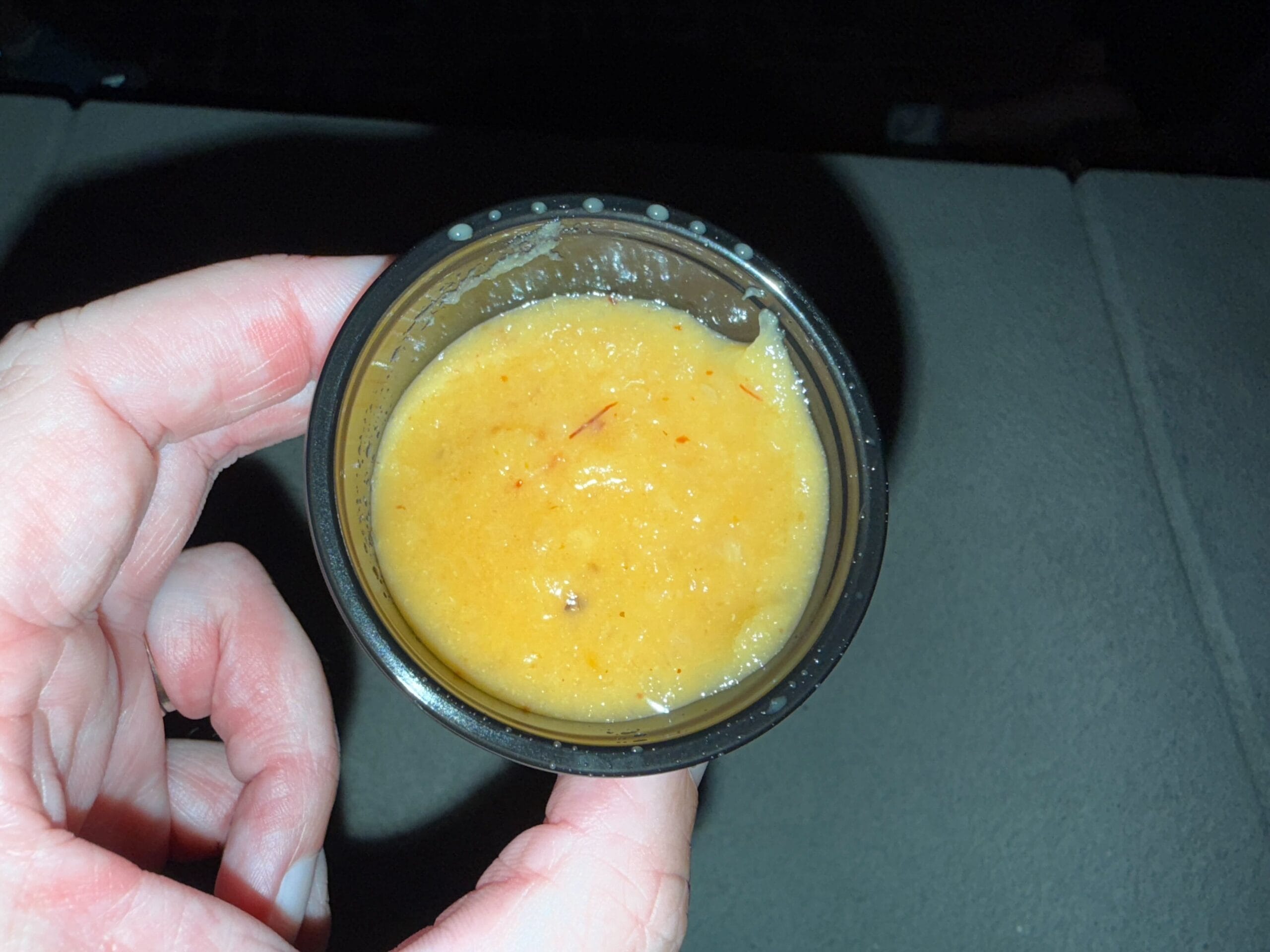 A hand holds a small black cup of thick, yellow-orange sauce—perfect for food reviews under bright light.