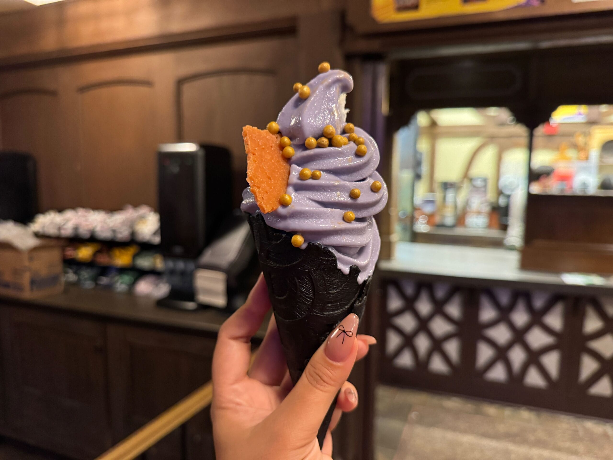 A hand holds a black cone with purple soft serve and gold sprinkles, perfect for food reviews in a rustic shop.