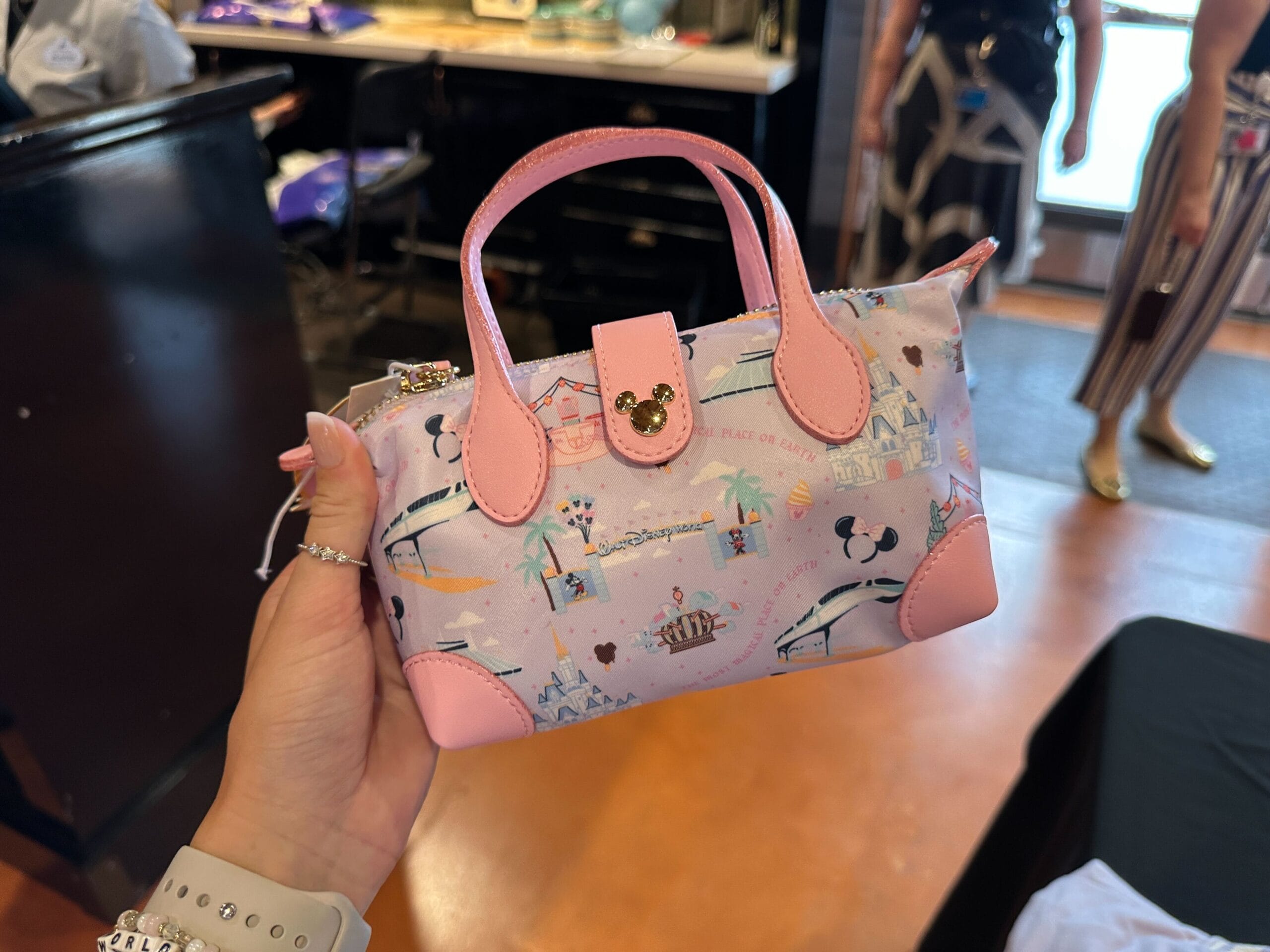 A hand holds a small pastel handbag with pink handles and Disney-themed illustrations, indoors.