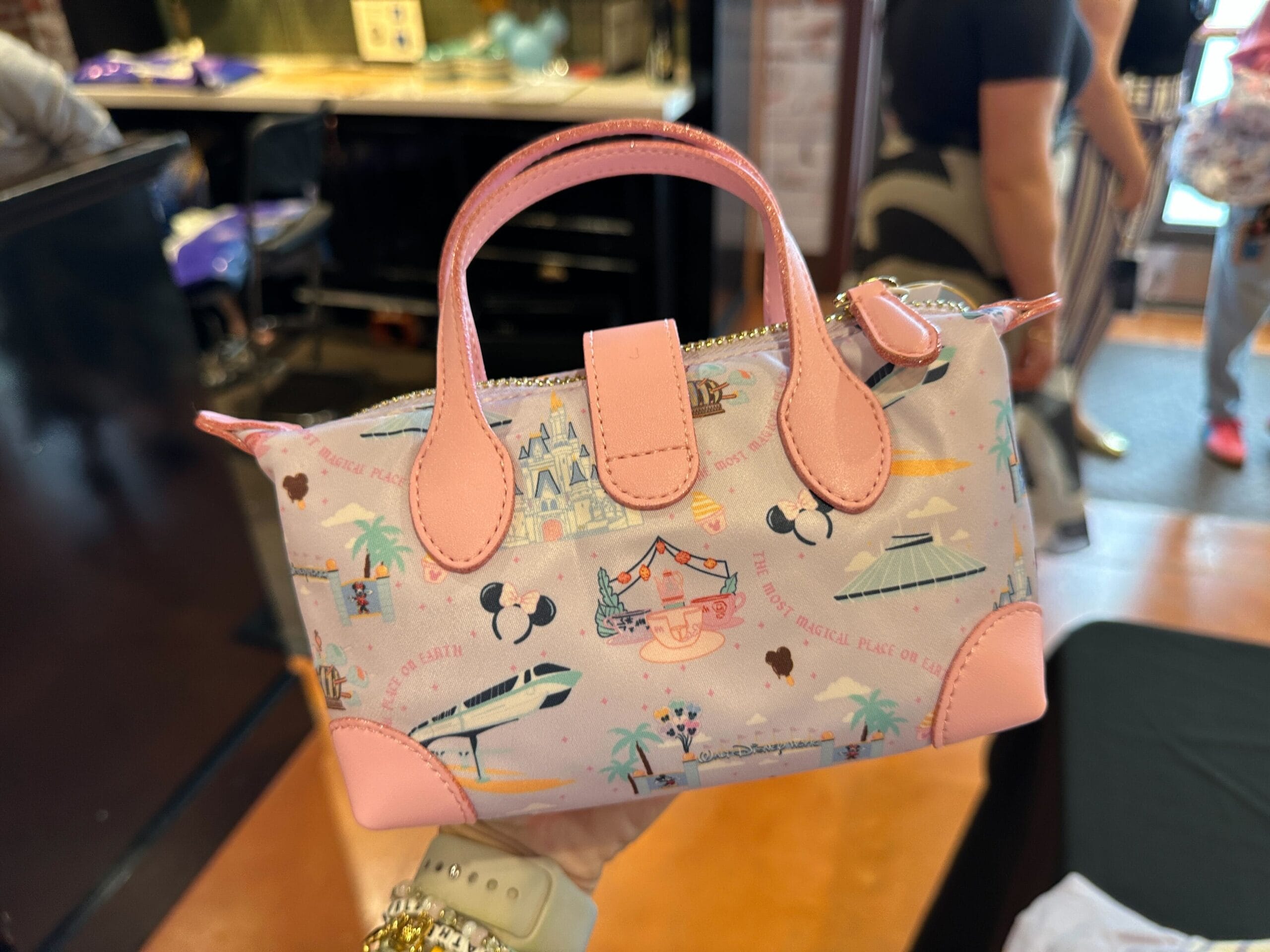 A small pastel pink handbag with Disney park icons, held up indoors by a person wearing a gold bracelet.