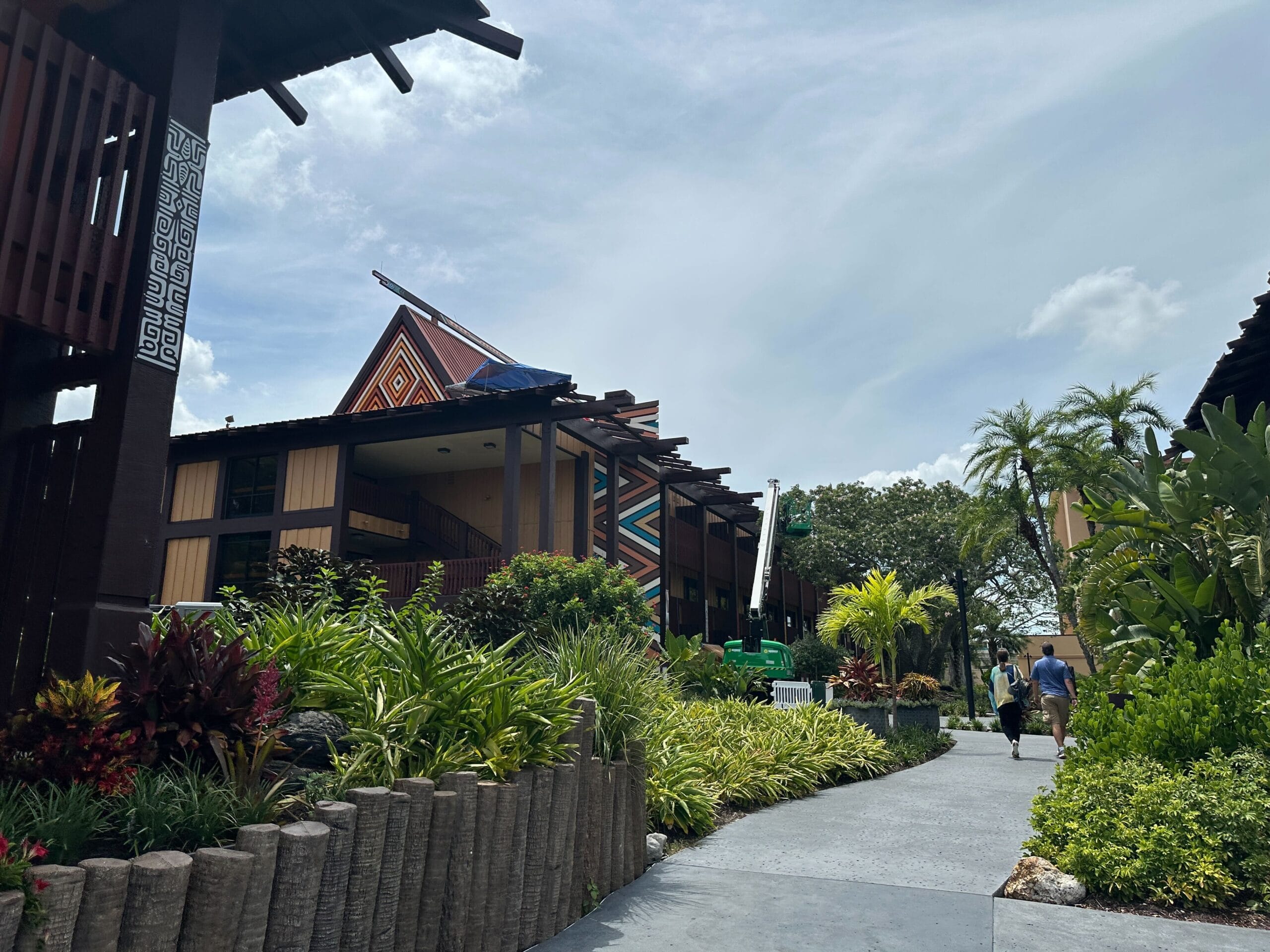 A tropical resort pathway with lush plants, wooden buildings, and two people walking under a partly cloudy sky.