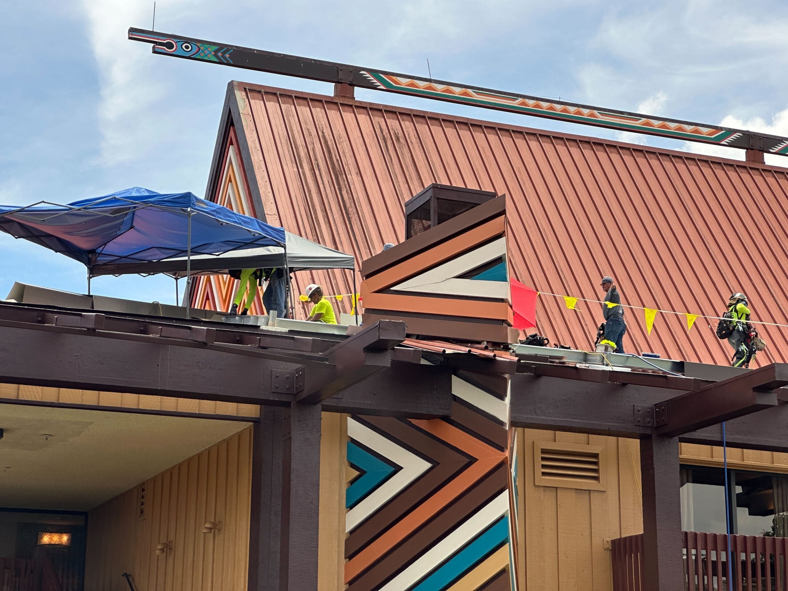 Workers in safety gear are on a slanted red roof near a decorated column with bold geometric patterns.