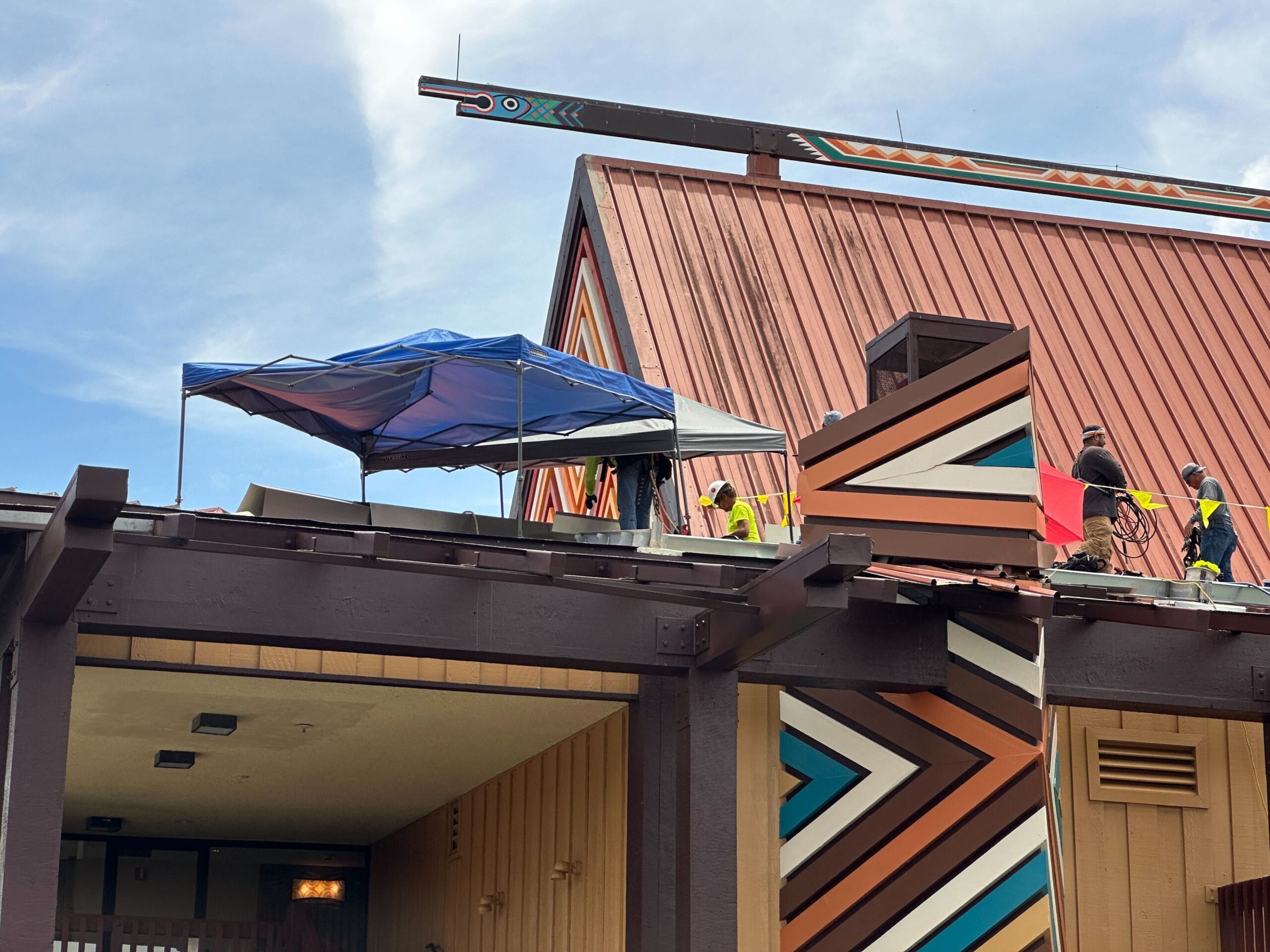 Construction workers repair a building roof with geometric patterns, working under a blue canopy on a sunny day.