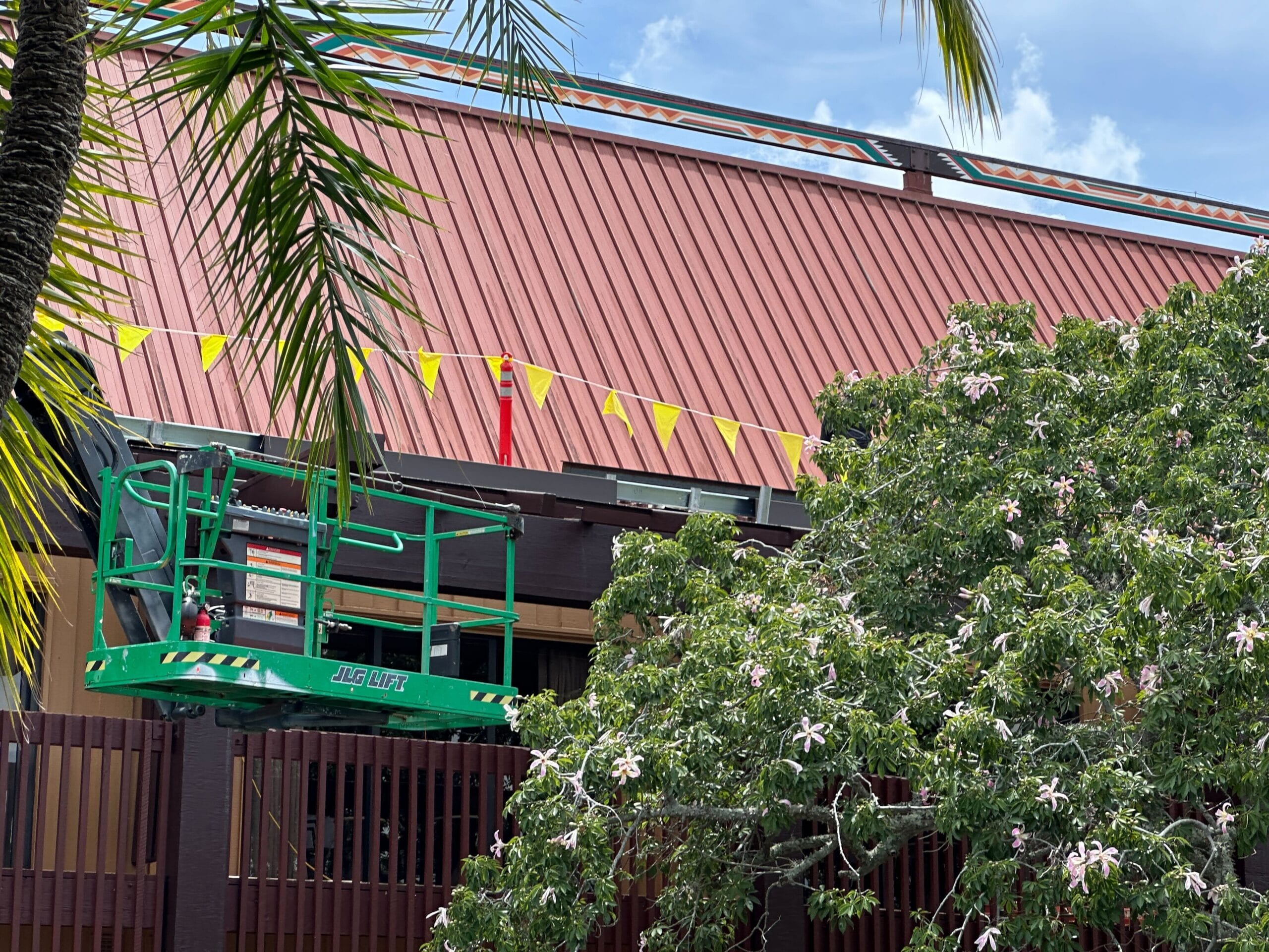A green lift near a brown building with yellow flags, palm leaves, and a flowering tree in the foreground.