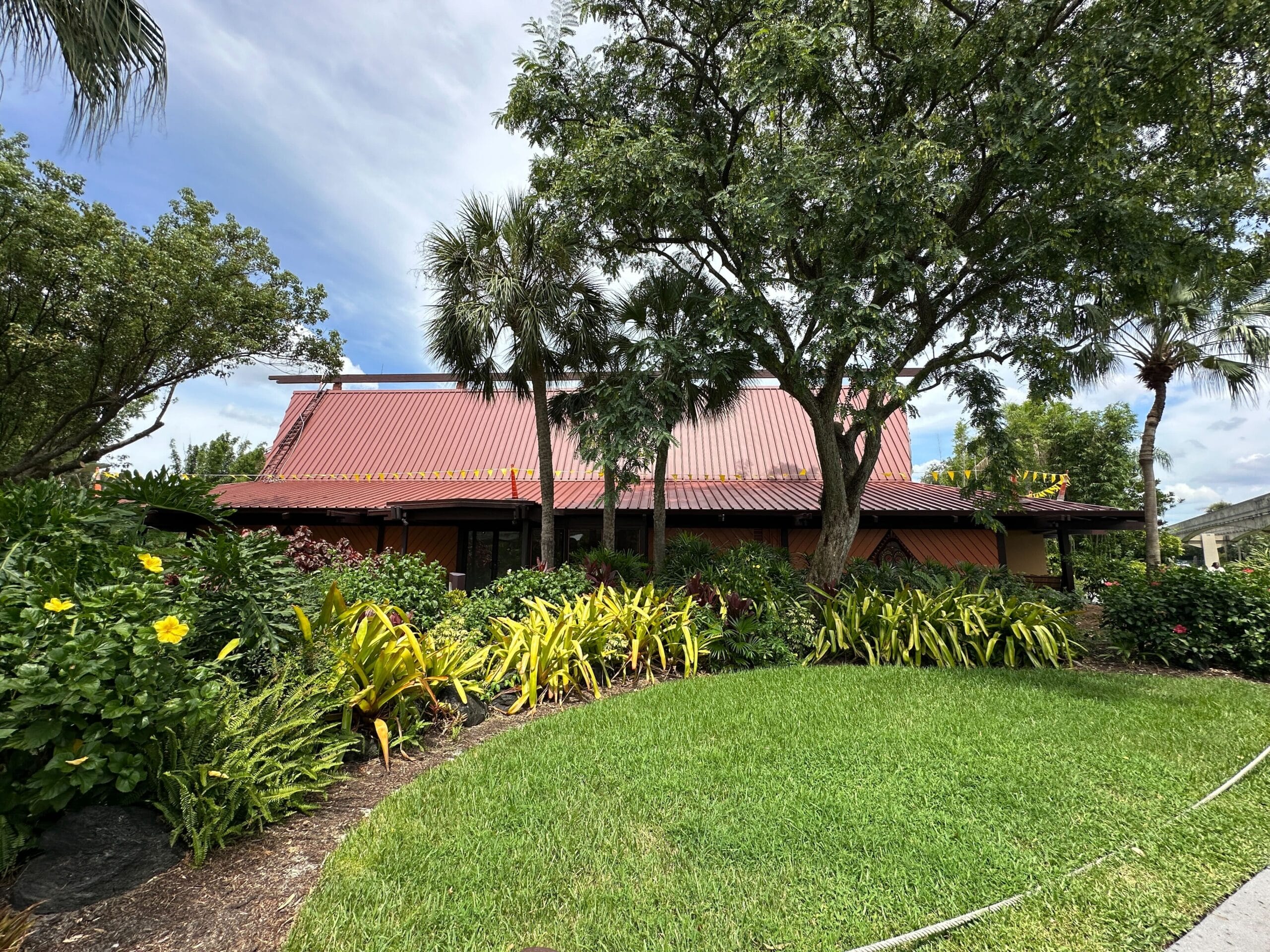 Restaurant with a red roof surrounded by lush green plants, trees, and a well-kept grassy lawn under a blue sky.