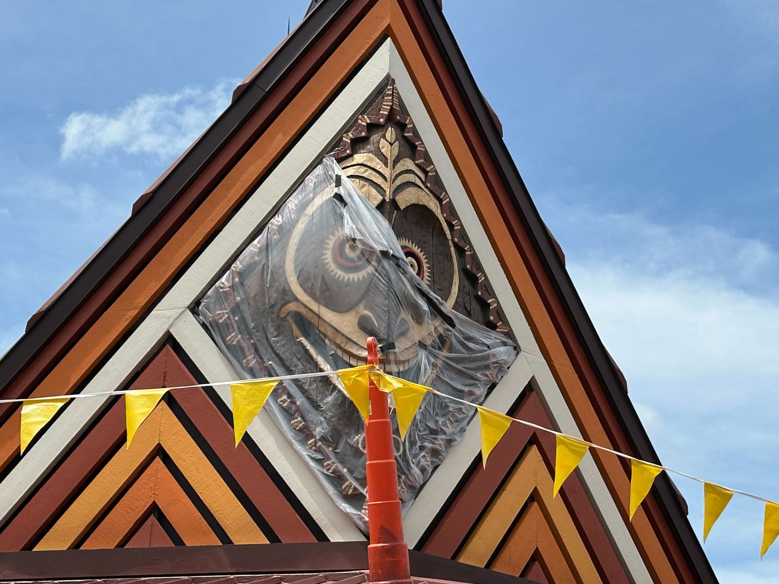 A colorful temple roof with a decorative owl detail and yellow triangular flags strung in front under a blue sky.