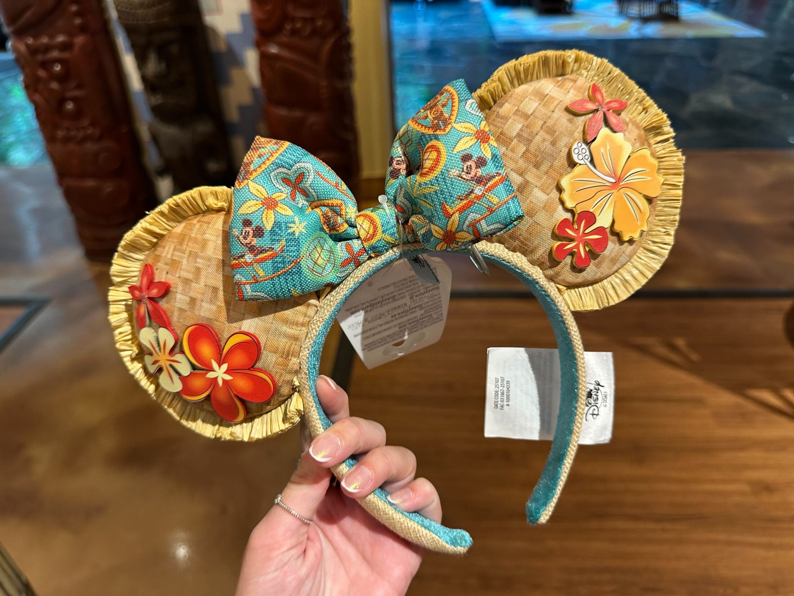 A hand holds a tropical-themed Mickey ears headband with floral designs and a colorful bow.