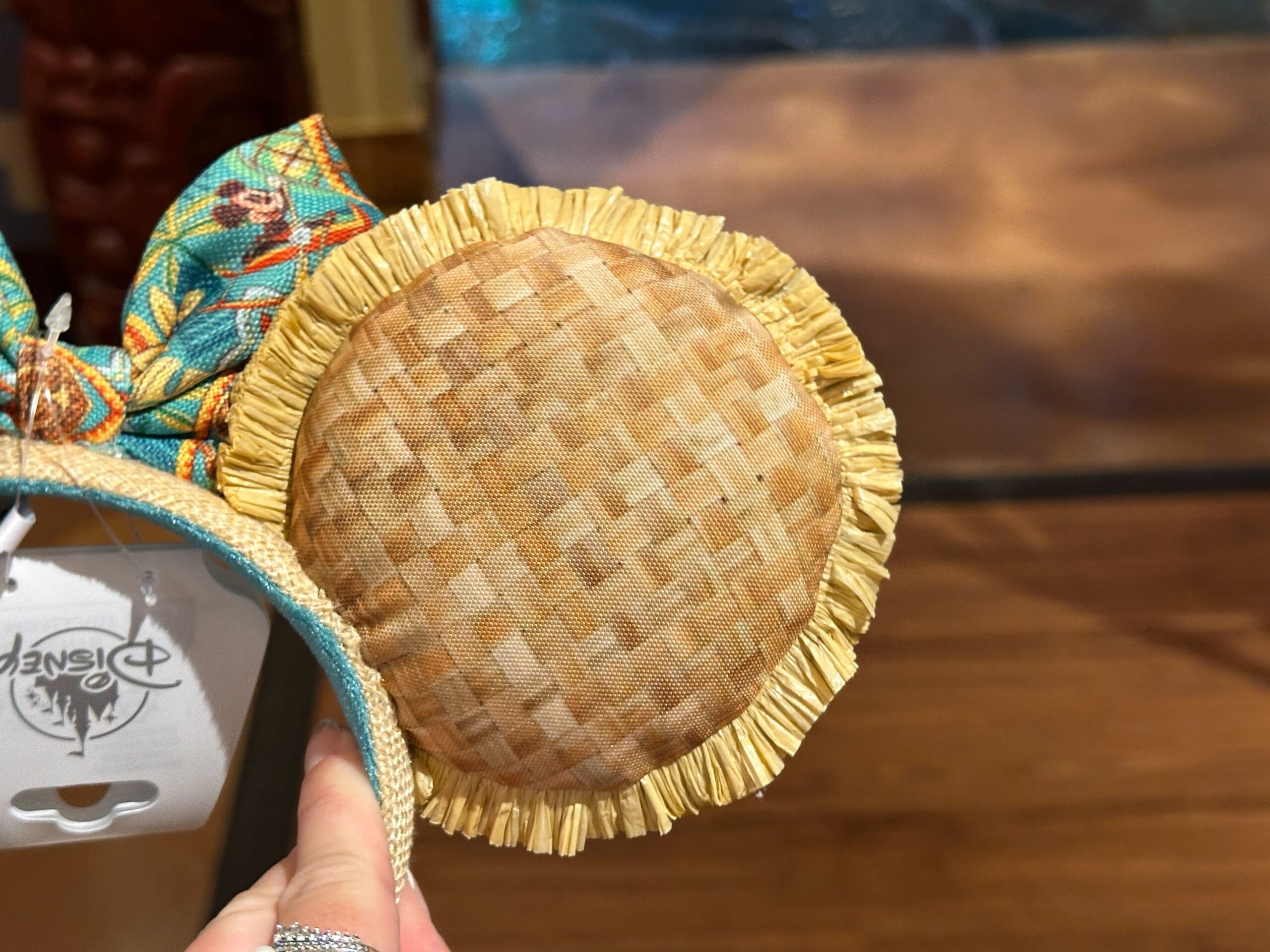 A hand holds a Disney headband with a round woven straw detail and yellow fringe trim against a wooden background.