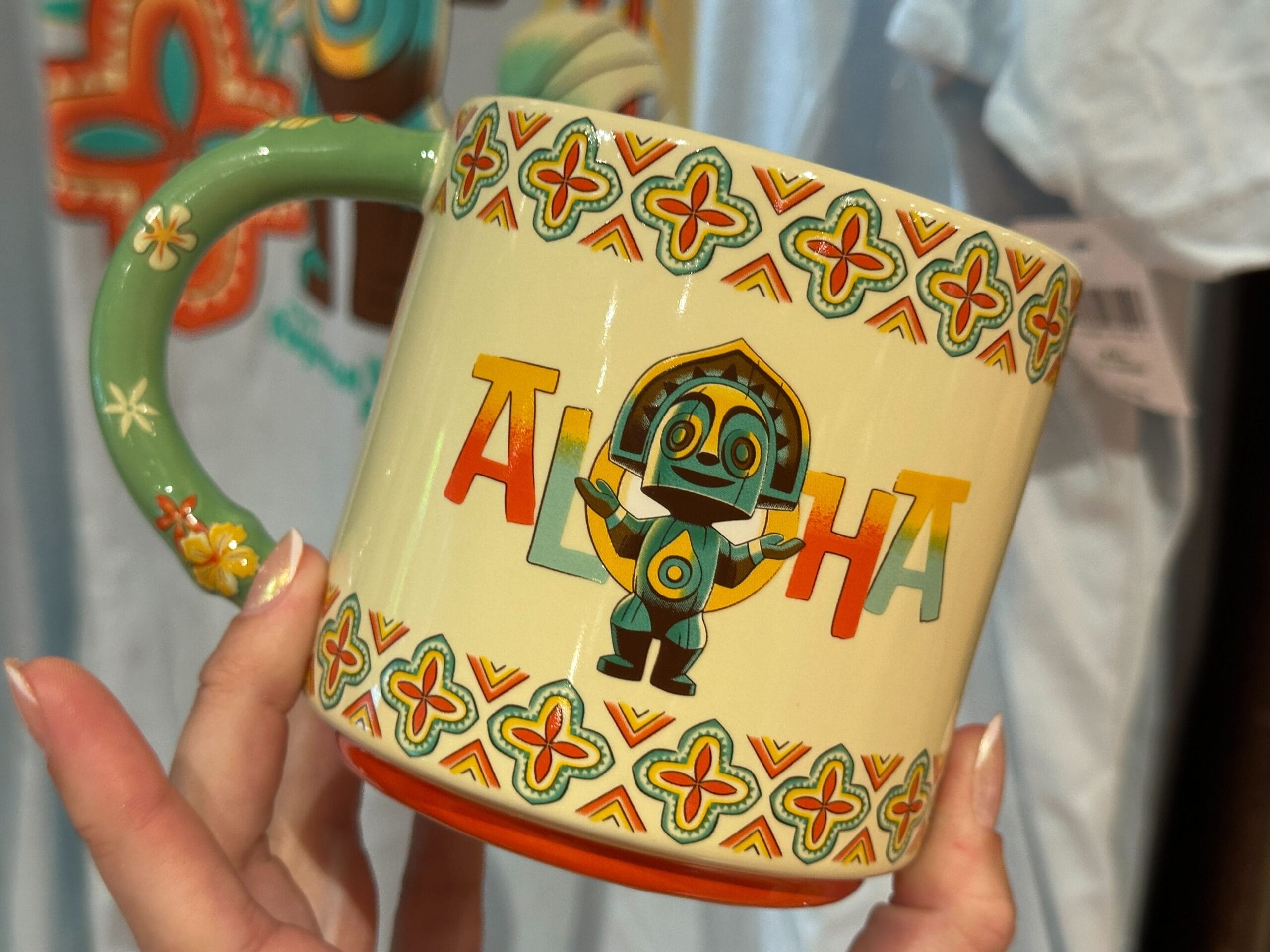 Hand holding a colorful mug with a tiki figure and the word "Aloha" surrounded by patterned designs.