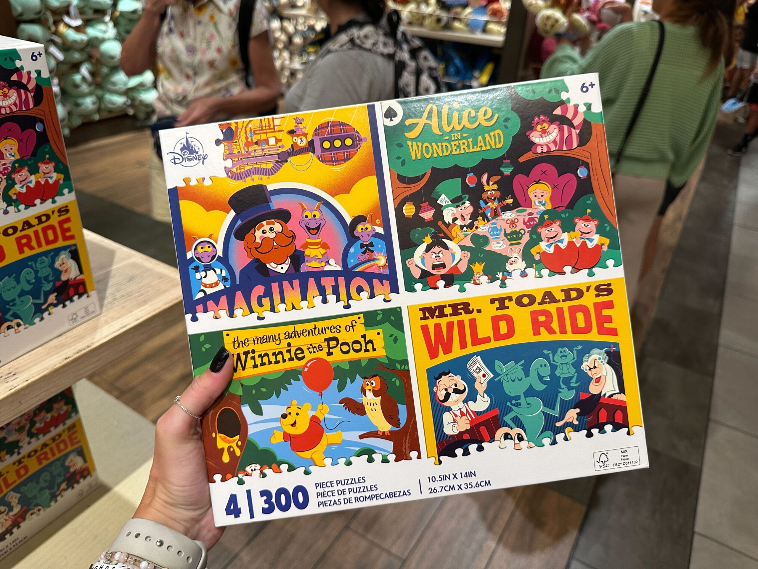 A hand holds a 300-piece Disney puzzle featuring Alice in Wonderland, Winnie the Pooh, Imagination, and Mr. Toad’s Wild Ride.
