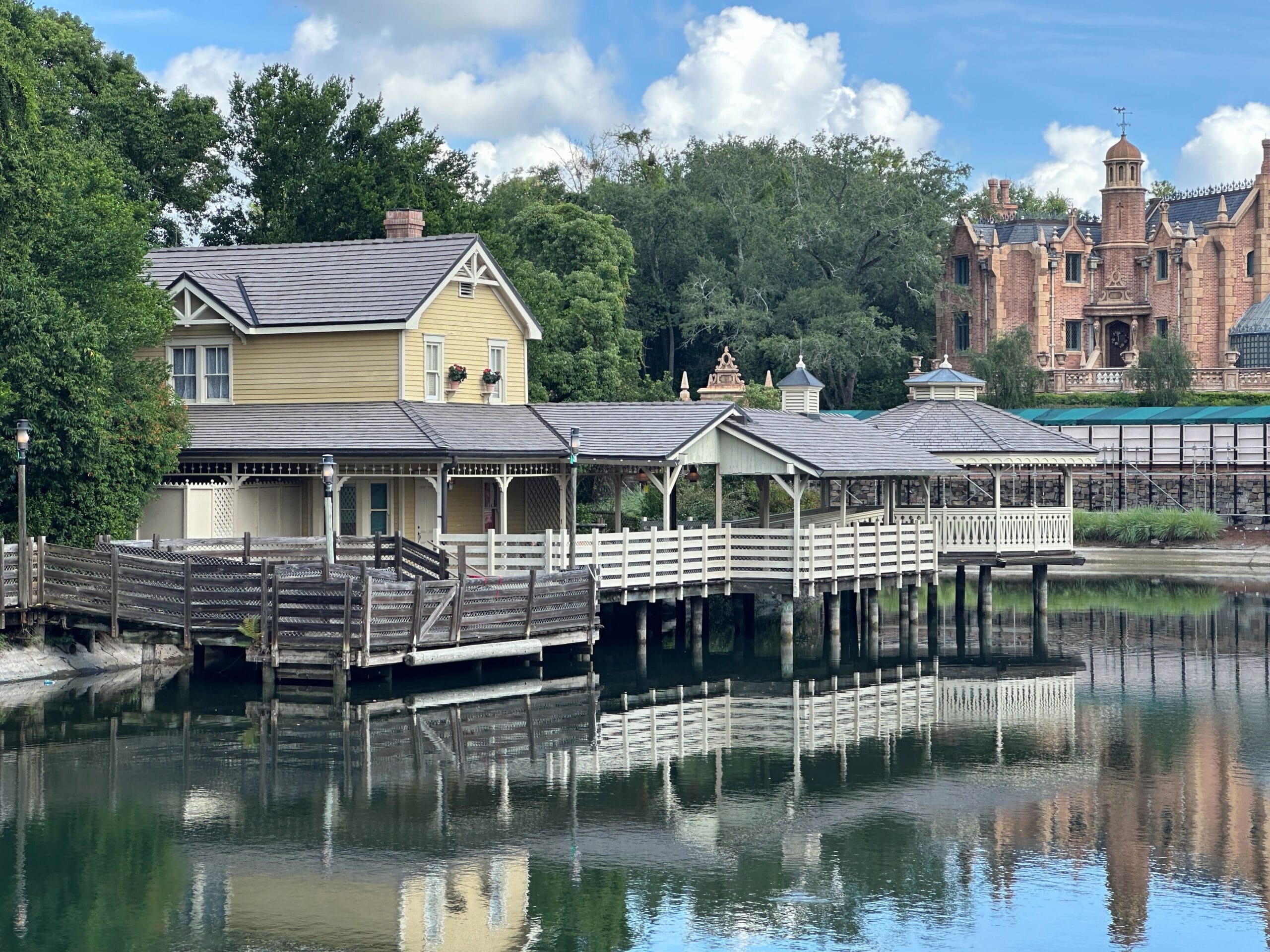 A yellow house and covered dock are reflected in a calm lake, with a large brick mansion in the background.