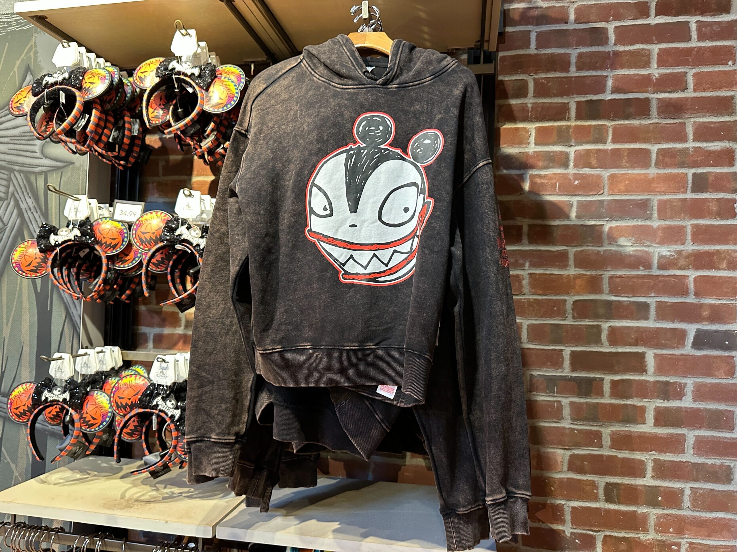 A black hoodie with a cartoonish monster face joins 2025 Disney Halloween Merch on a rack beside festive headbands.