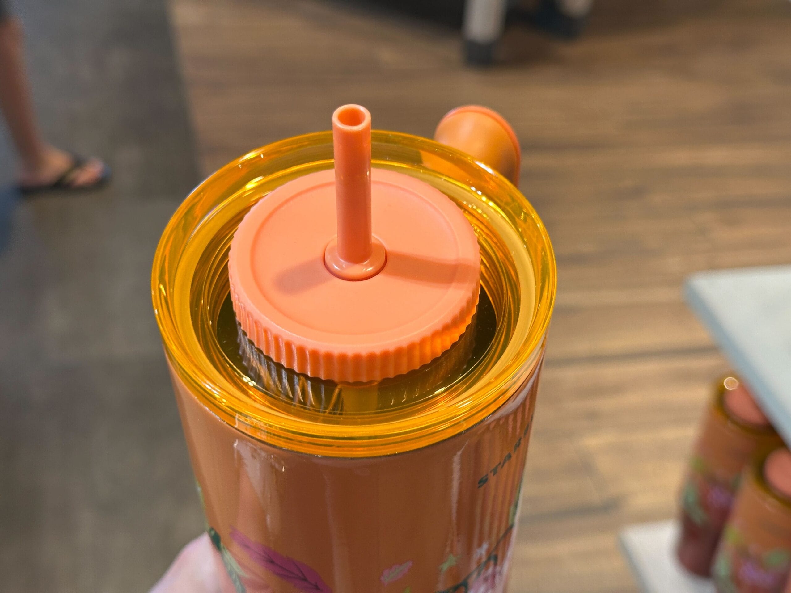 Close-up of a hand holding a Starbucks orange tumbler with lid and straw, set against a wooden floor background.