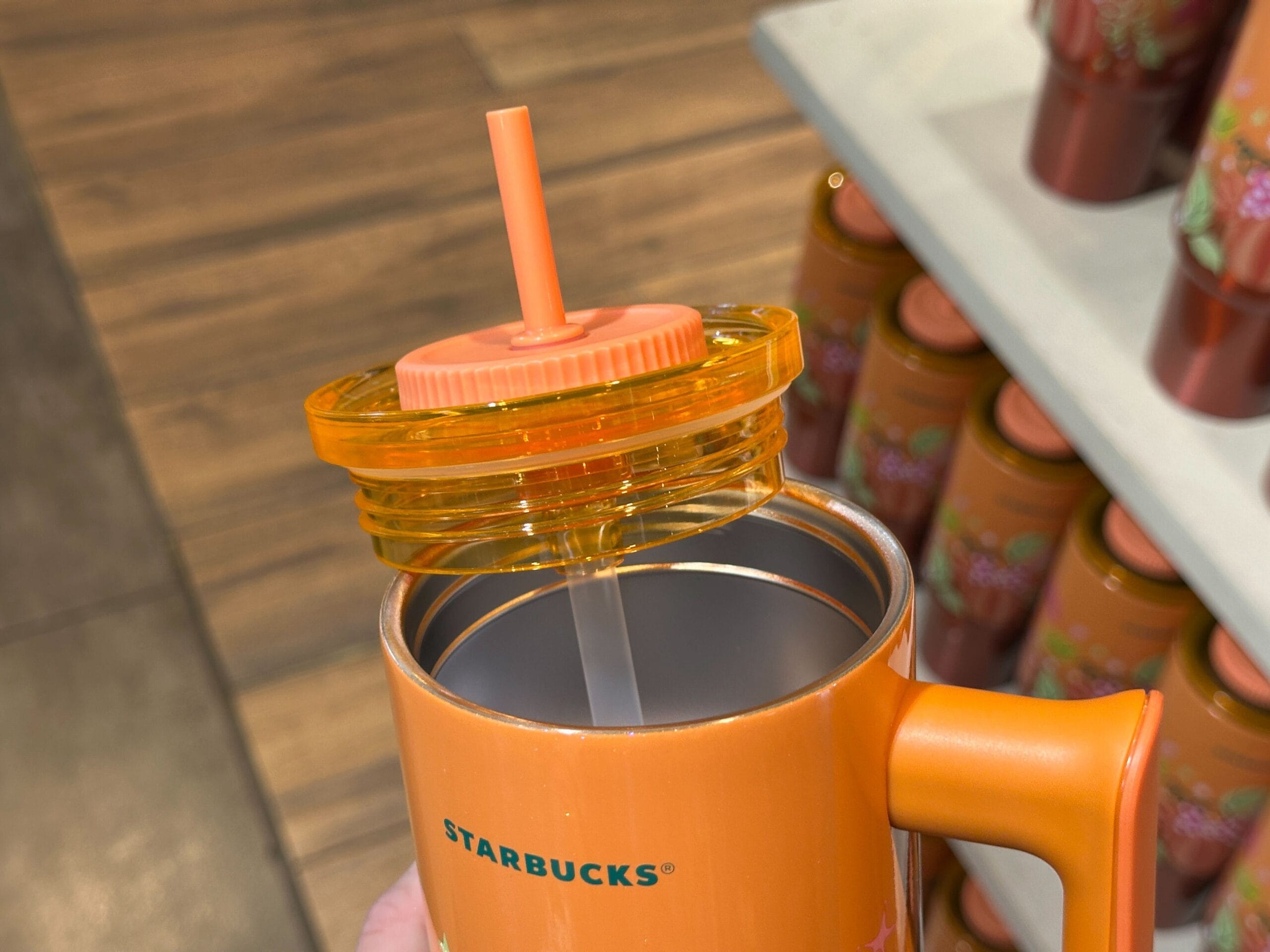 A hand holds an orange Pumpkin Spice Starbucks mug with lid and straw, surrounded by similar mugs on a store display.