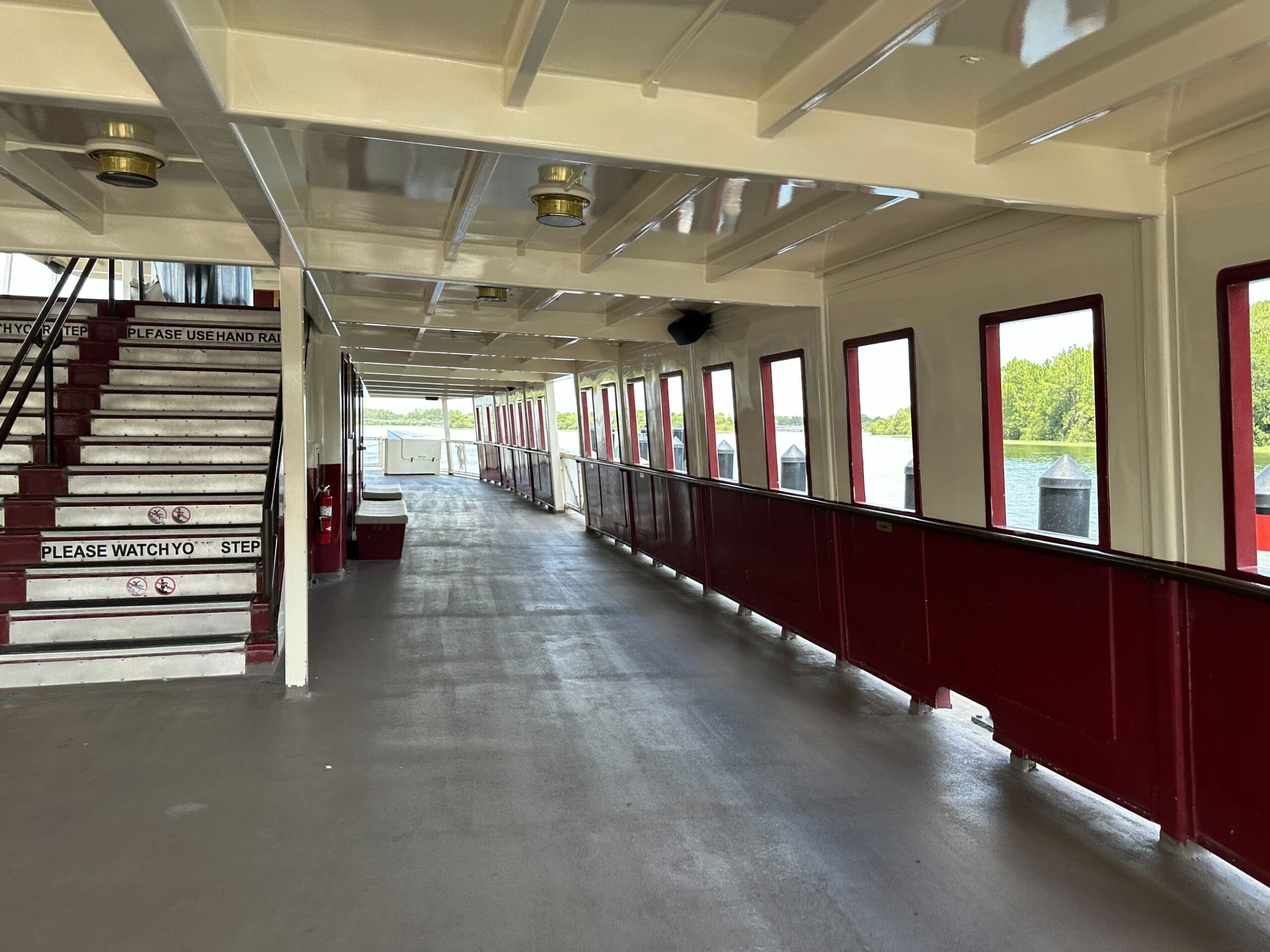Empty ferry deck with maroon railings, windows showing water and trees, and stairs marked “Please Watch Your Step.”