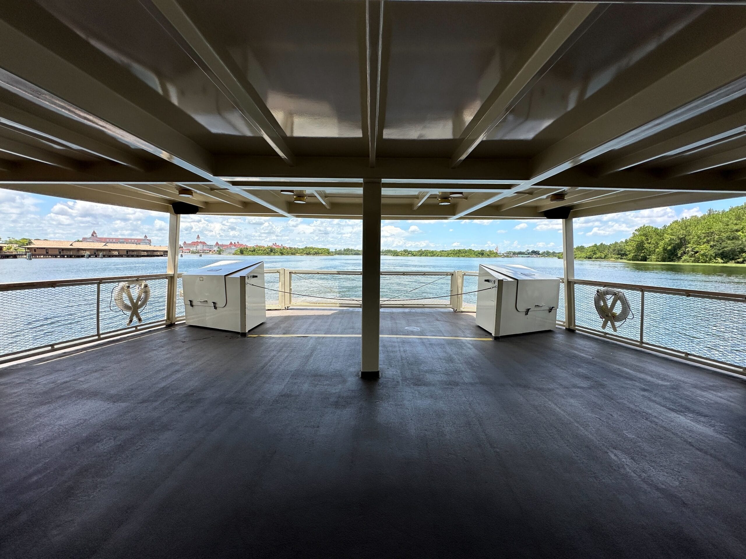 View from a covered ferry deck, looking out over water toward buildings and trees under a partly cloudy sky.