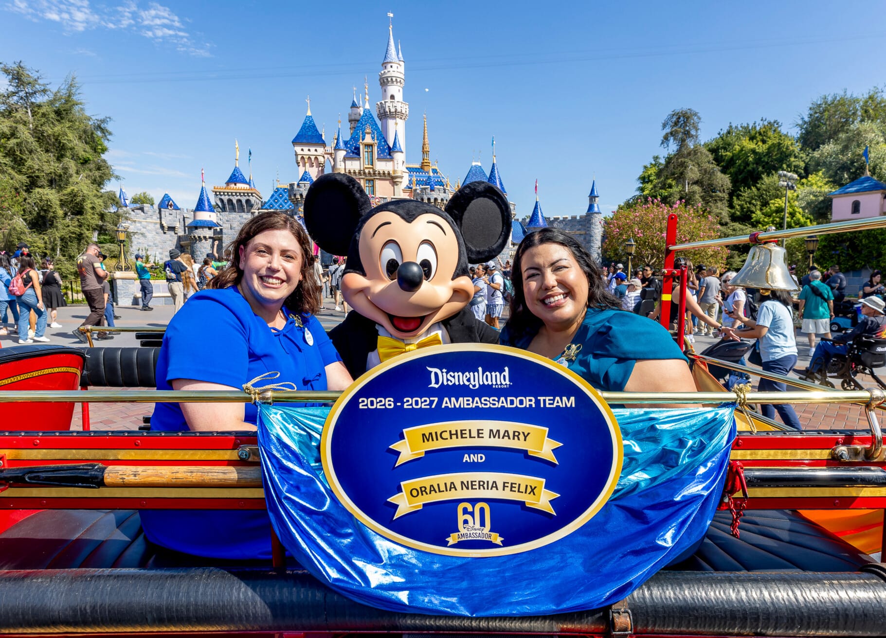Two women and Mickey Mouse pose in front of Disneyland's castle, sitting behind a Disneyland Ambassador Team banner.