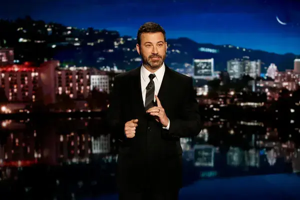 A man in a black suit speaks on stage at night with a cityscape, like on Jimmy Kimmel Live.