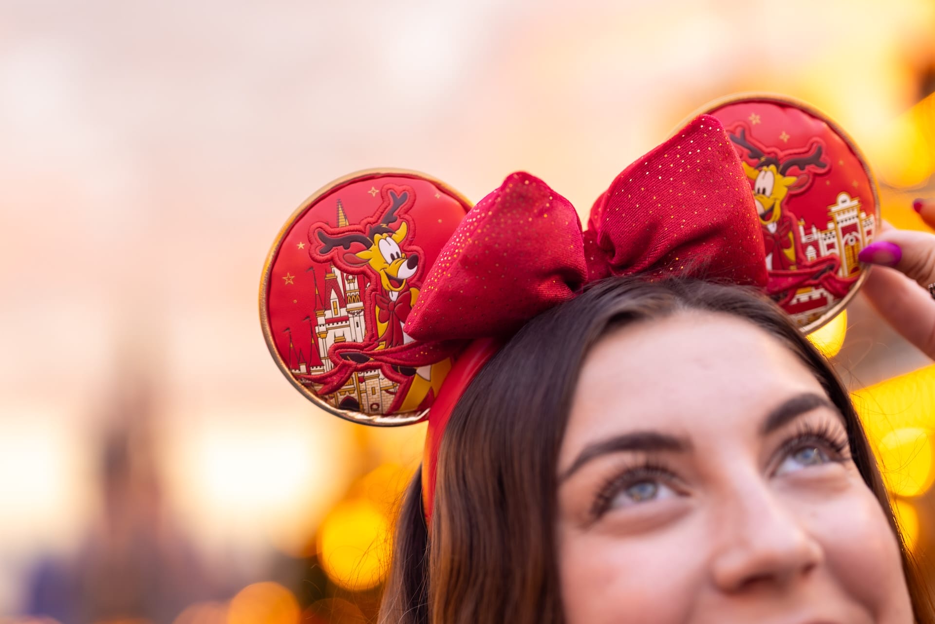 Person wearing red Minnie Mouse ears and Spirit Jersey, smiling with sunset and lights in the background.