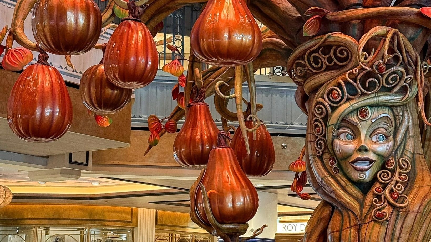 A whimsical Pumpkin Tree sculpture with orange gourds and a carved face brings Halloween charm to the shopping mall.