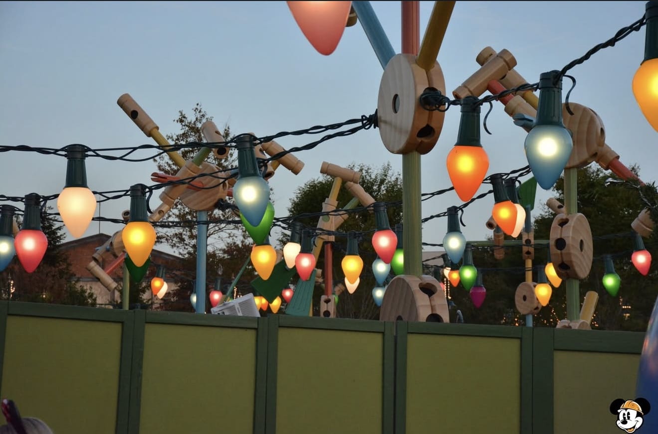 Colorful, oversized string lights hang above a green fence, with large toy-like decorations in the background at sunset.