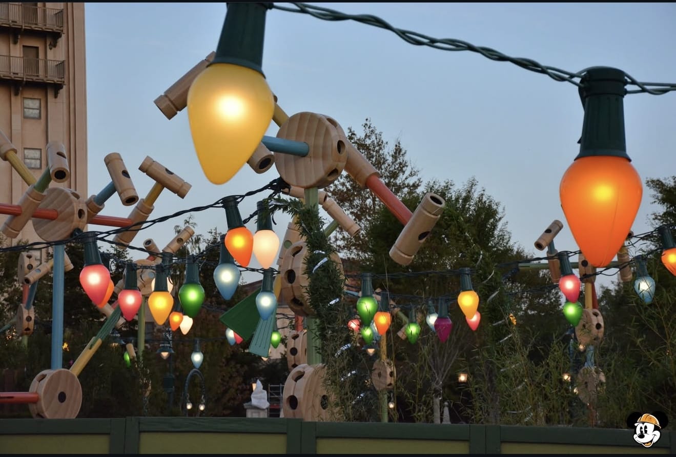 Colorful oversized Christmas lights strung above a themed outdoor area with tinker toy decorations and trees.
