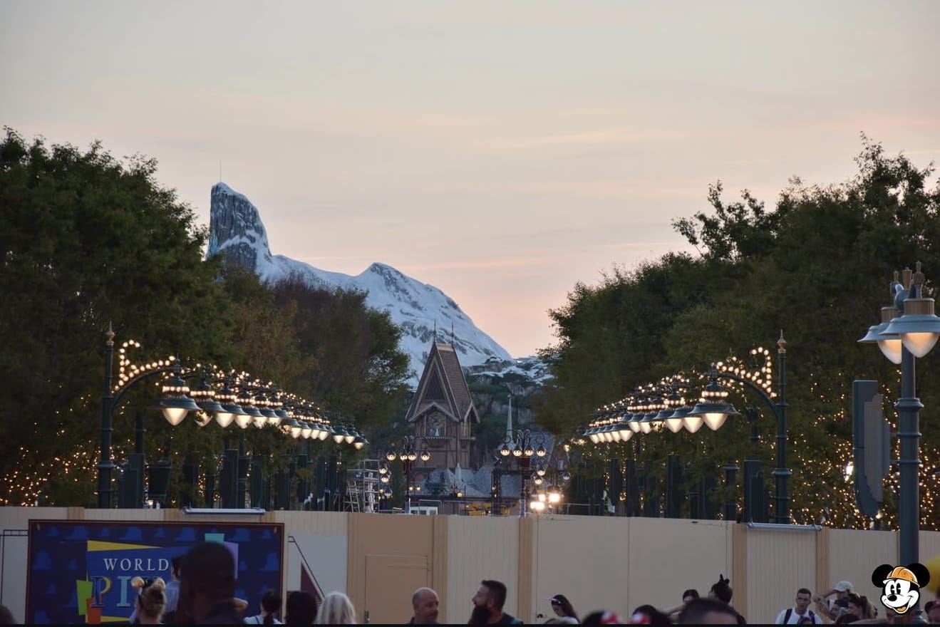 A view of a mountain and a castle with lit trees and lampposts lining a pathway at sunset, people walking below.