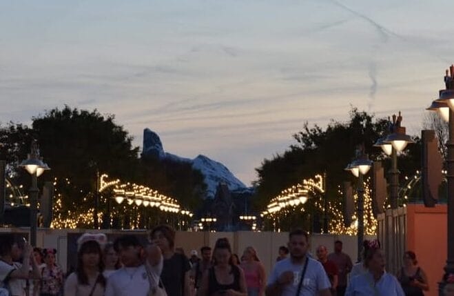 People walk along a lit street at dusk near neon signs and construction walls, with trees and mountains in the distance.