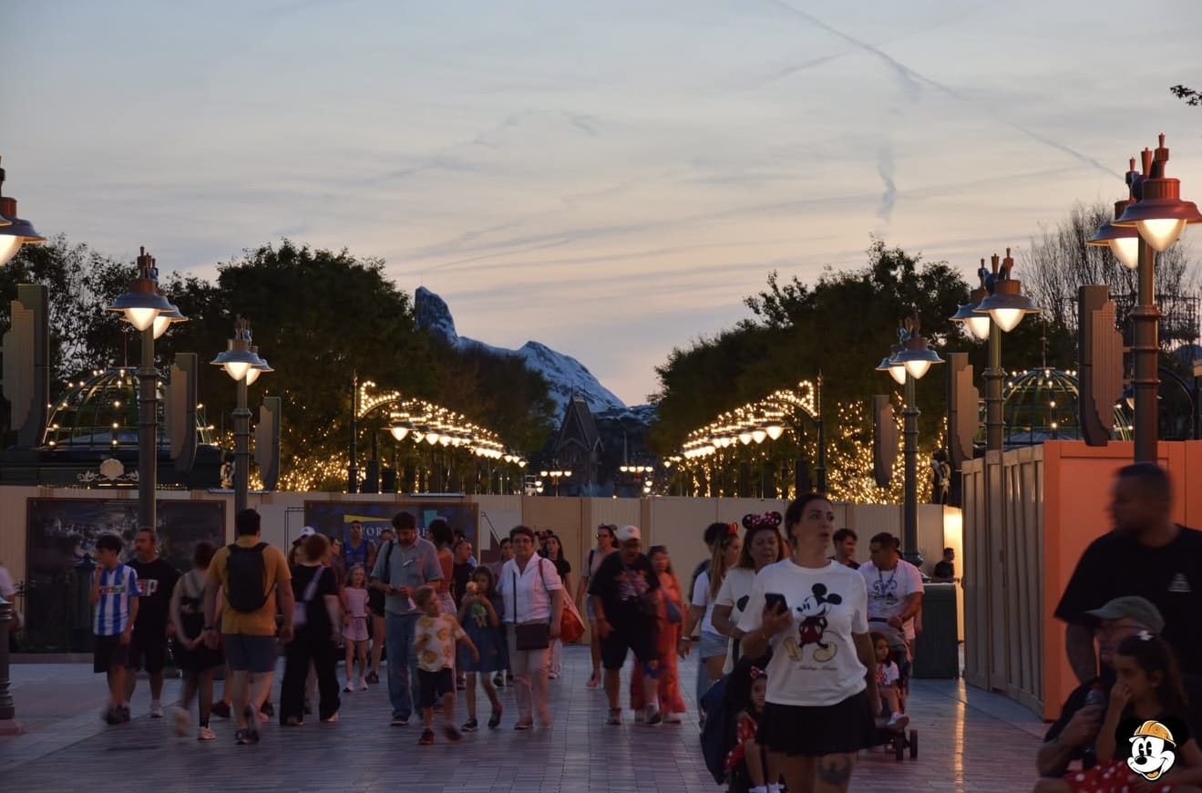 Crowds walk down a tree-lined avenue at sunset with lights and a mountain in the distance at Disneyland.
