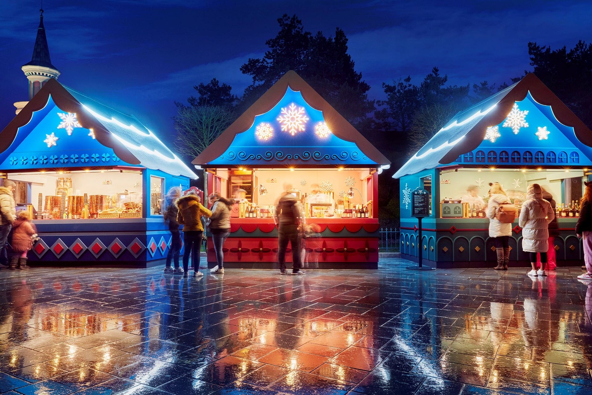 People visit brightly lit holiday market stalls at night, as if transported to magical Disneyland Paris under a dark blue sky.