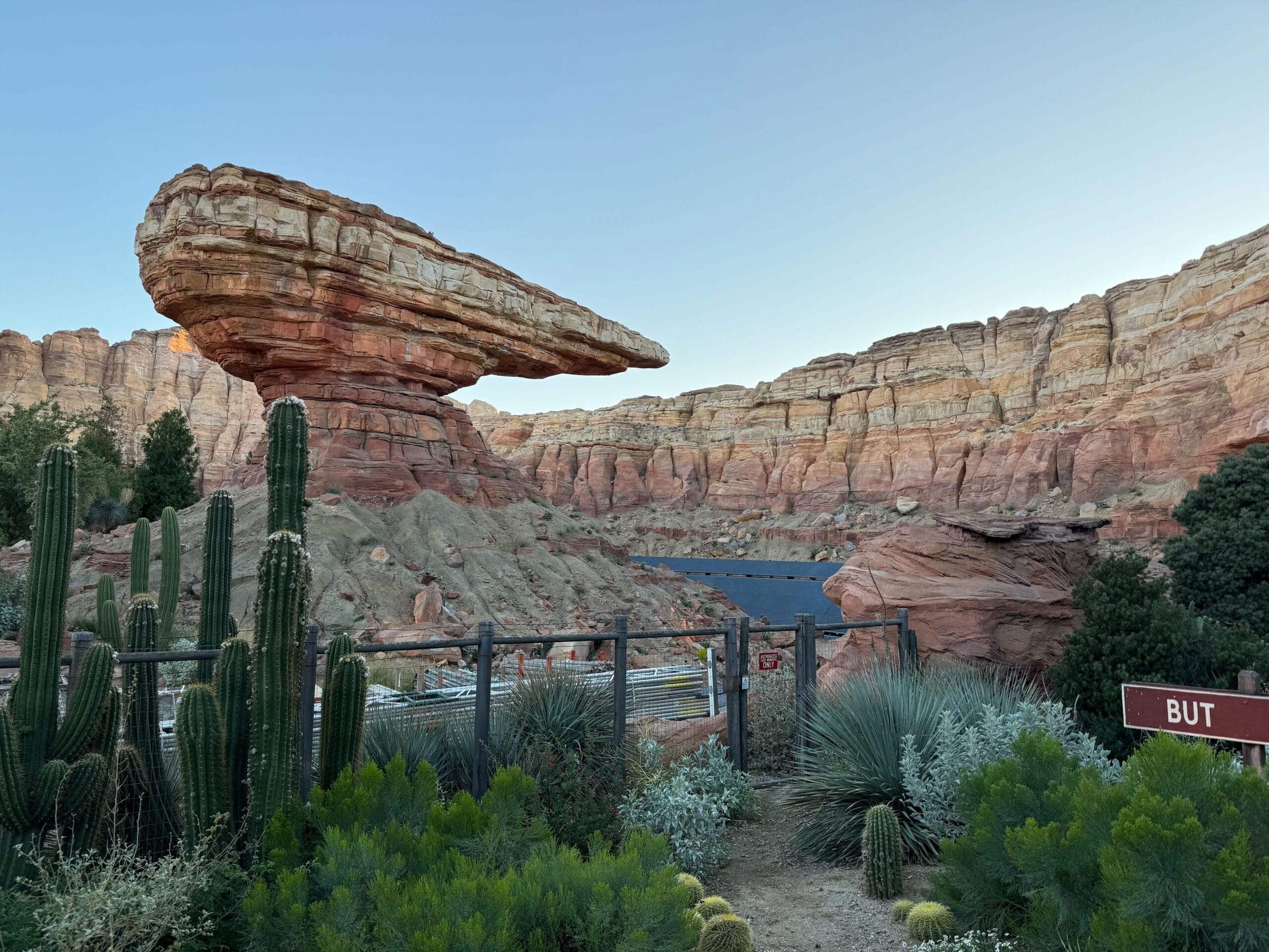 Red rock formations and desert plants with a fence and sign in the foreground, under a clear sky.