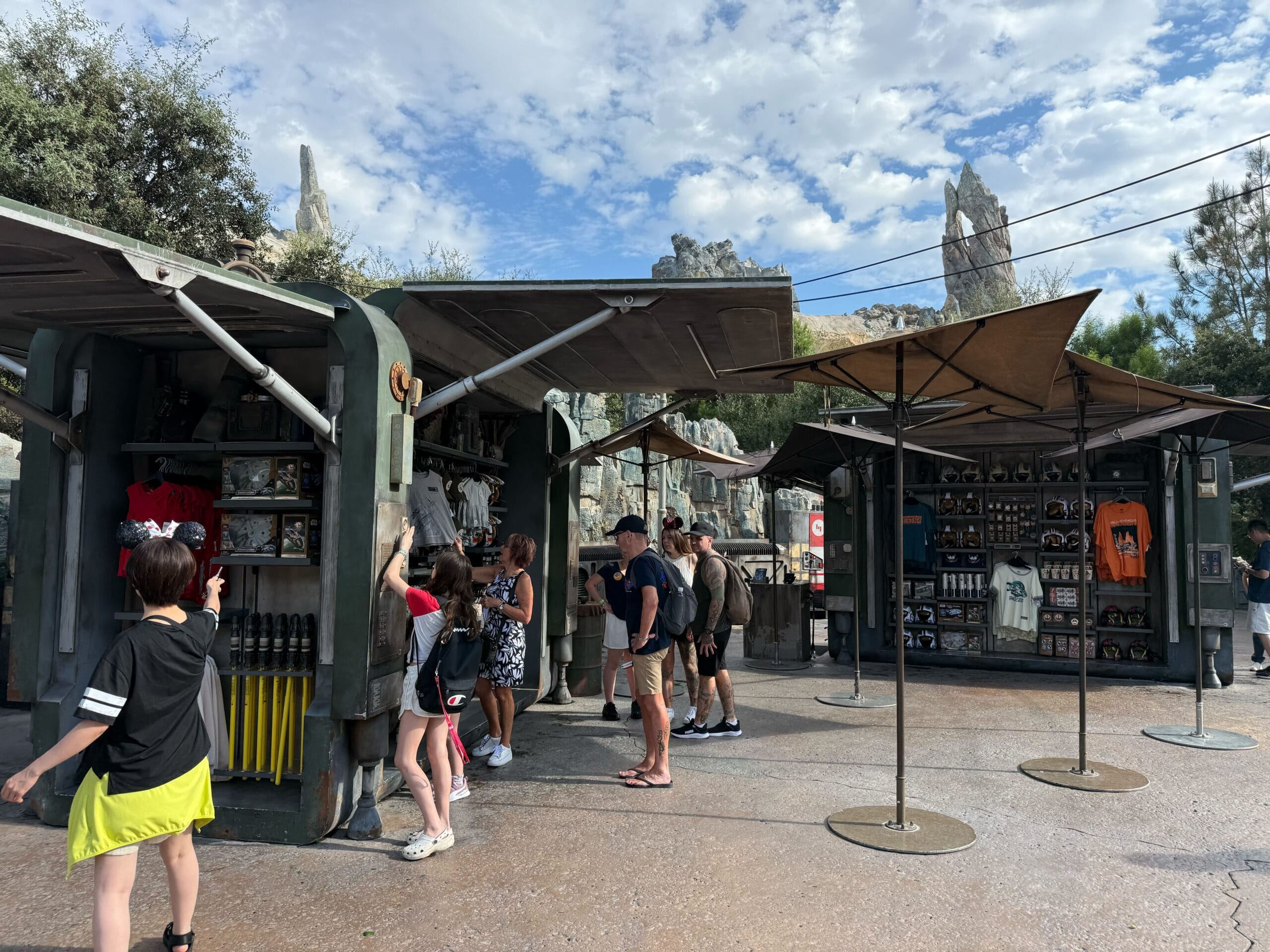 People browse Star Wars shirts at merchandise stalls with sci-fi decor and rocky spires in Disneyland.