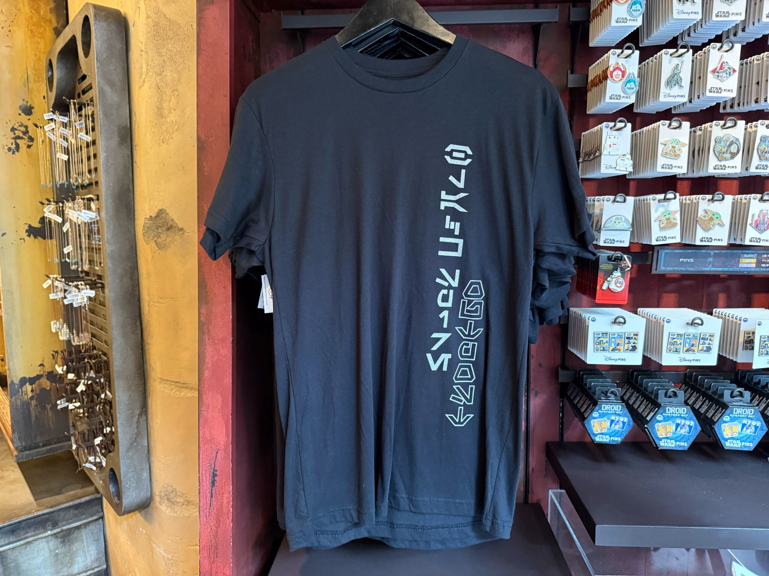 Black T-shirt with teal vertical alien script hangs in a store display among other Star Wars shirts.