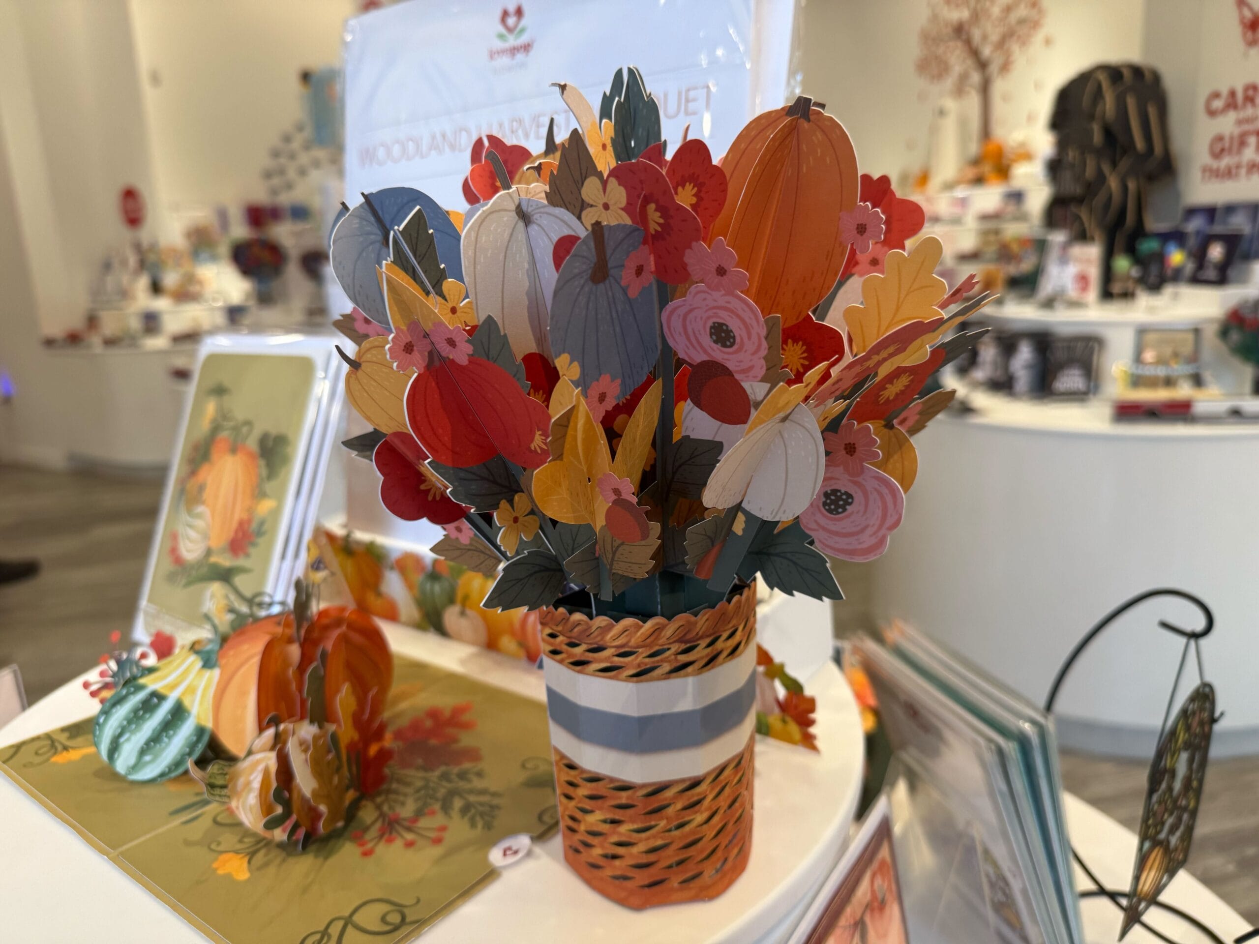 A colorful paper bouquet with pumpkins and flowers in a striped vase, displayed on a store counter.