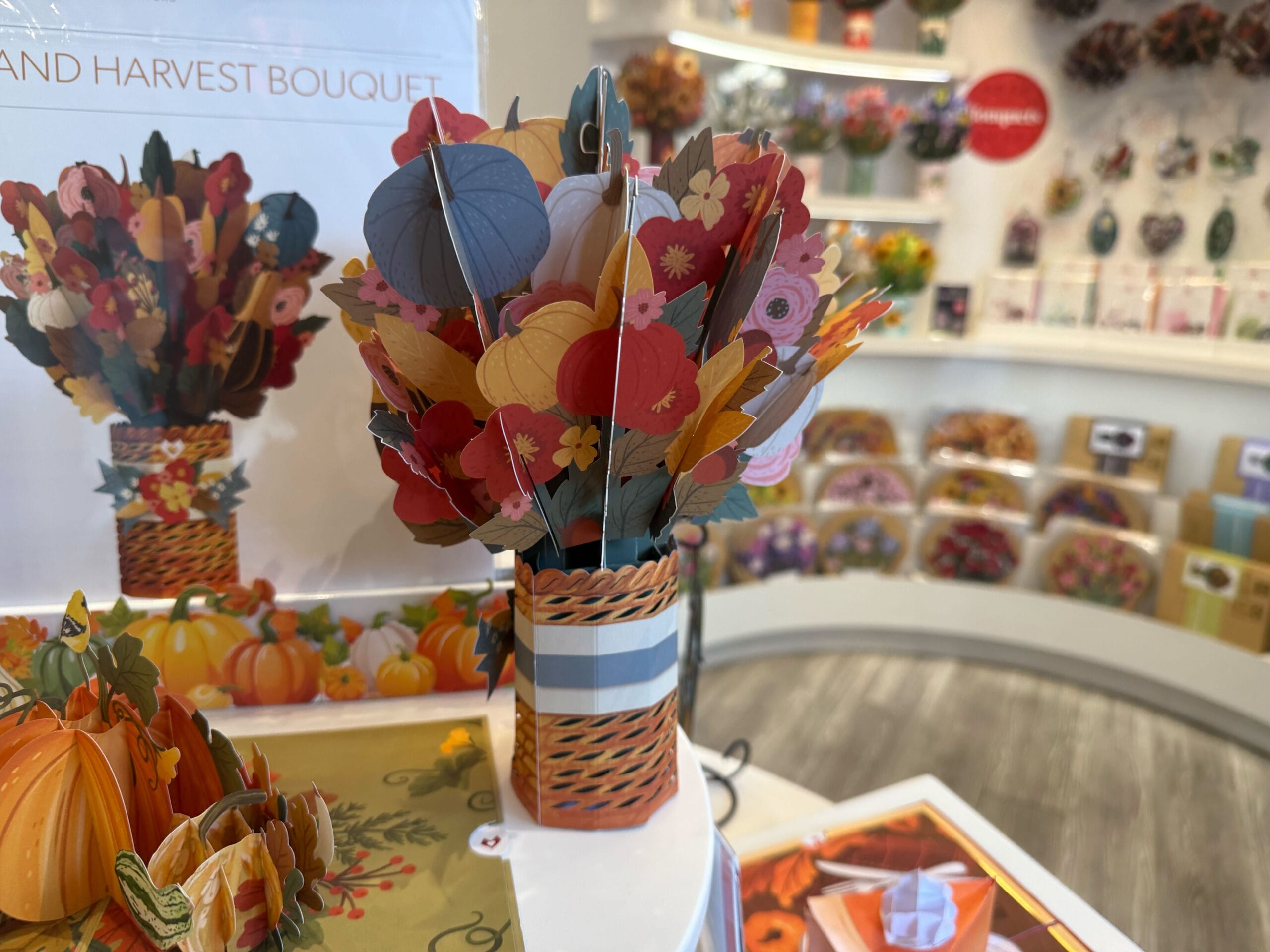 A colorful paper bouquet in a striped vase on display in a gift shop with cards and decorations in the background.