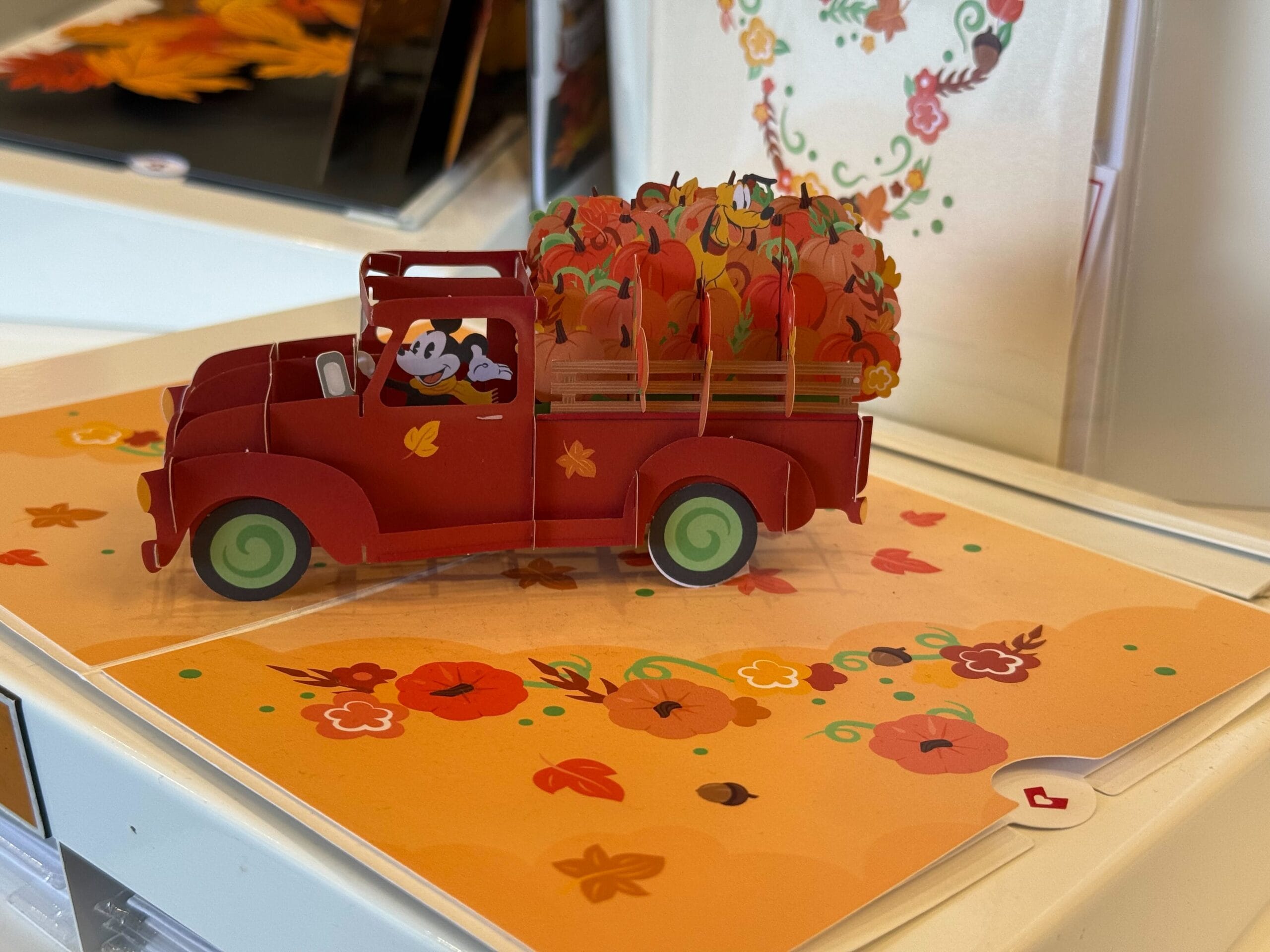 A red pop-up truck card filled with pumpkins sits on an orange surface decorated with fall leaves and flowers.