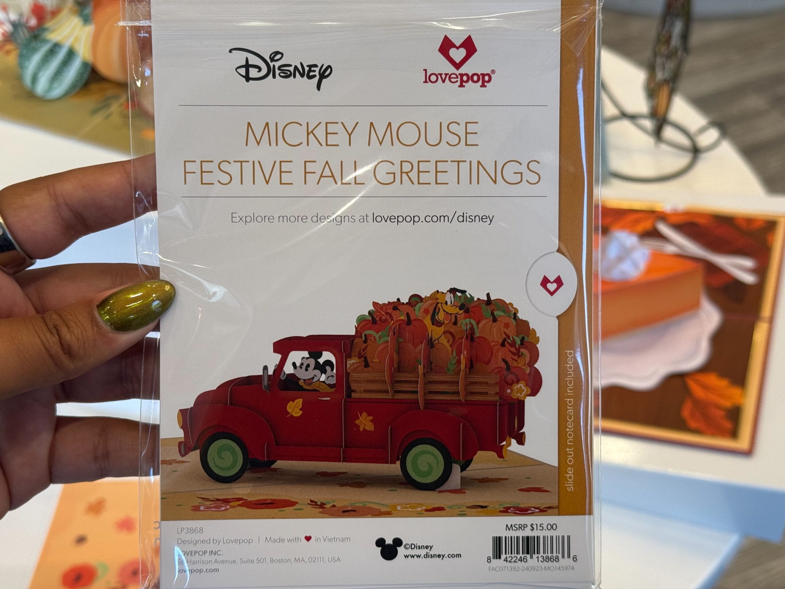 Hand holding a Lovepop card featuring Mickey Mouse driving a red truck filled with pumpkins, with fall decor in the background.
